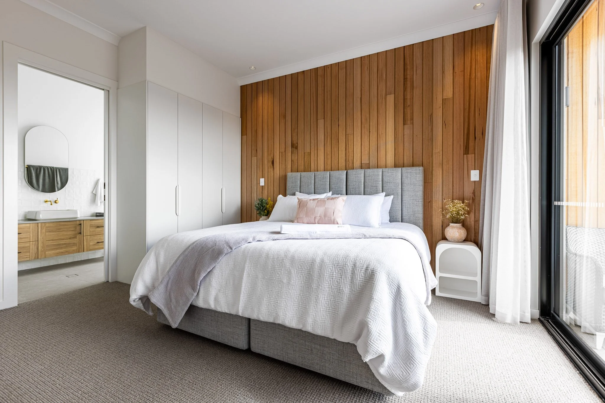 A modern bedroom with a bed featuring a gray upholstered headboard, white bedding, and decorative pillows, next to a white nightstand with a small plant, with a wooden accent wall and large window with white curtains.