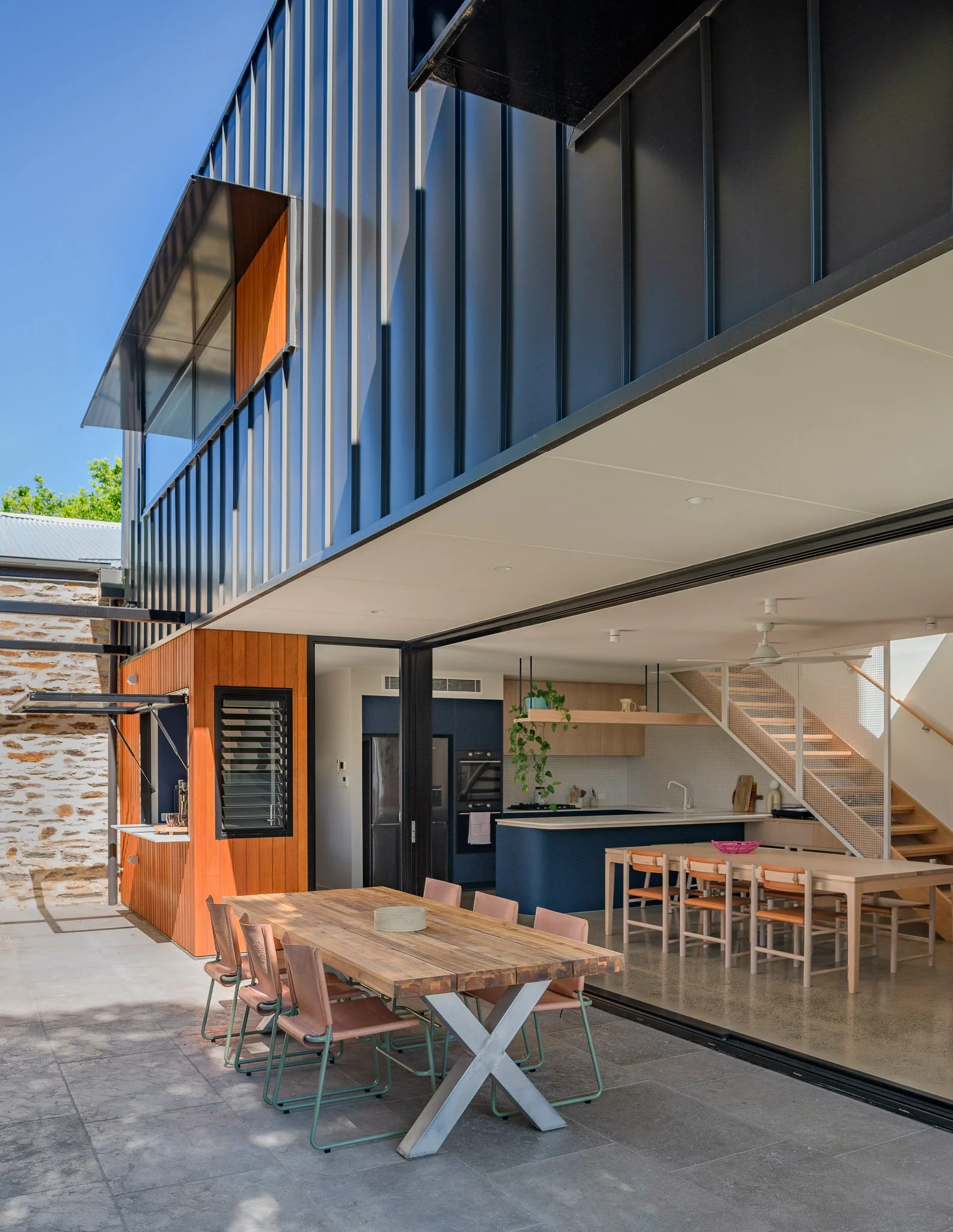 Modern outdoor dining area with a large wooden table and pink chairs on a concrete patio, adjacent to a contemporary house with metal siding, large windows, and an open kitchen visible through a sliding glass door.