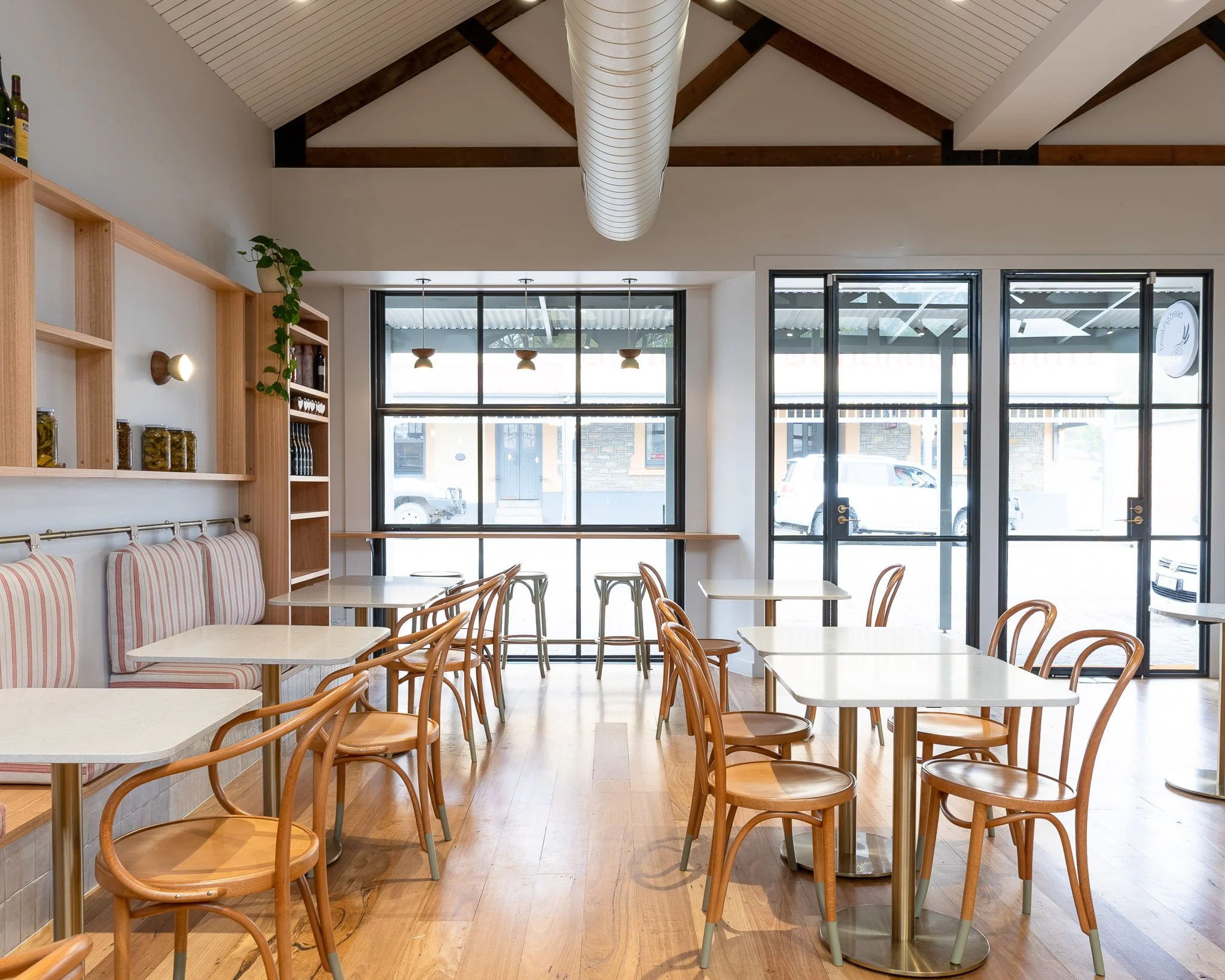 Inside of a modern cafe with light wood furniture, including tables and chairs, and large windows allowing natural light, with a ceiling featuring exposed wooden beams and a large central ventilation duct.