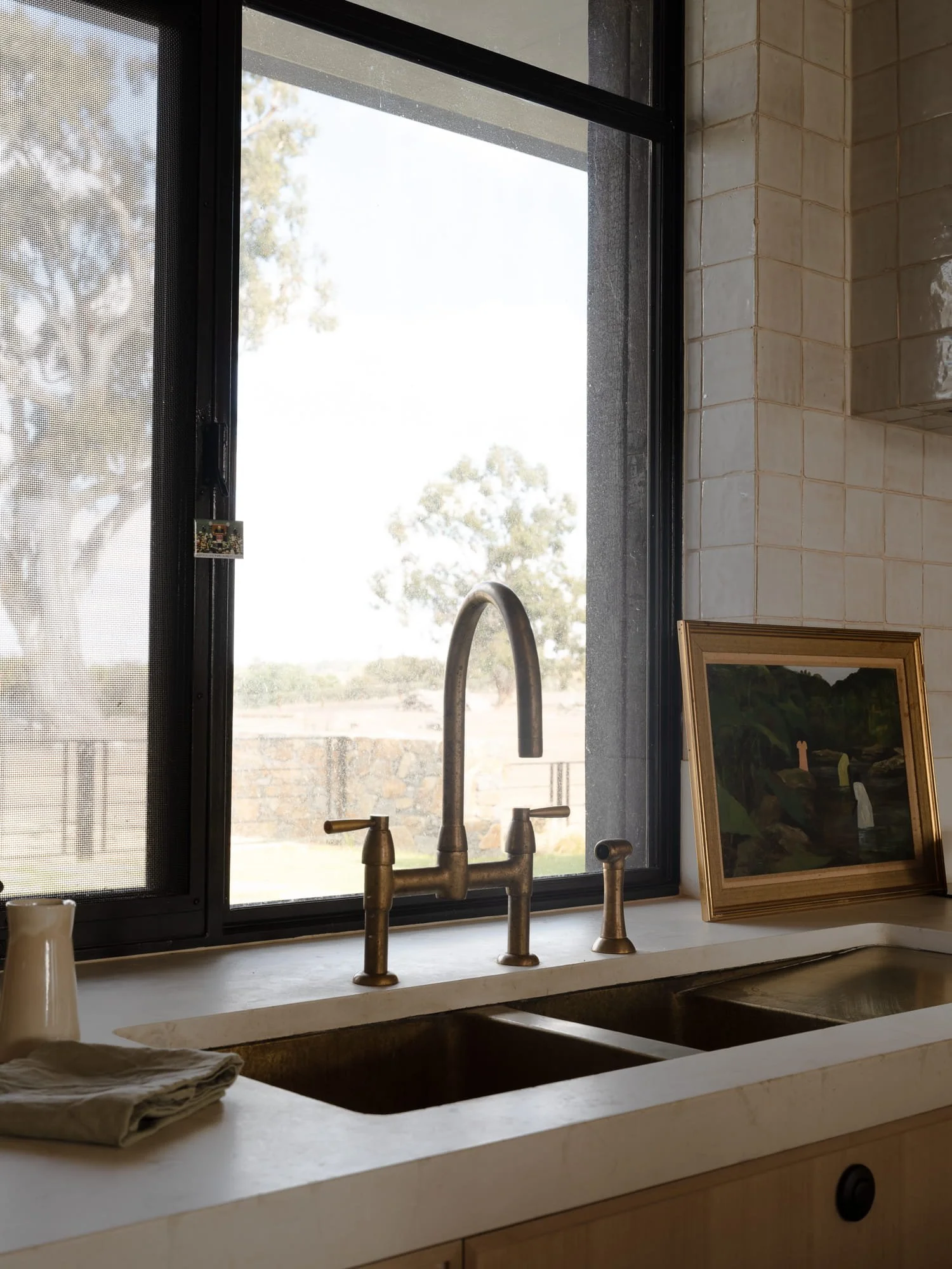 View of a kitchen window with a faucet and a framed painting on the countertop. Outside, trees and clear sky are visible.