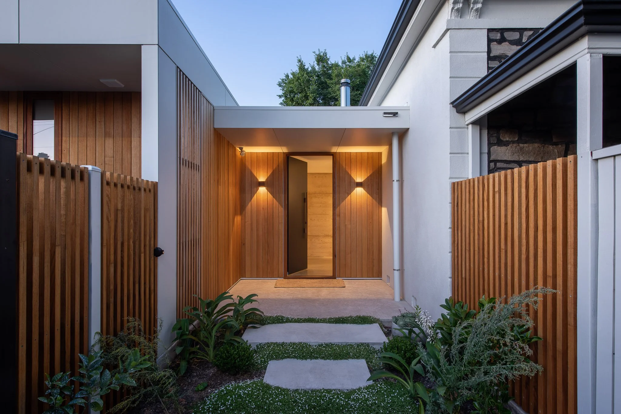 Entrance to a modern house with wooden fencing, a pathway, and garden plants, illuminated by outdoor lighting at dusk.