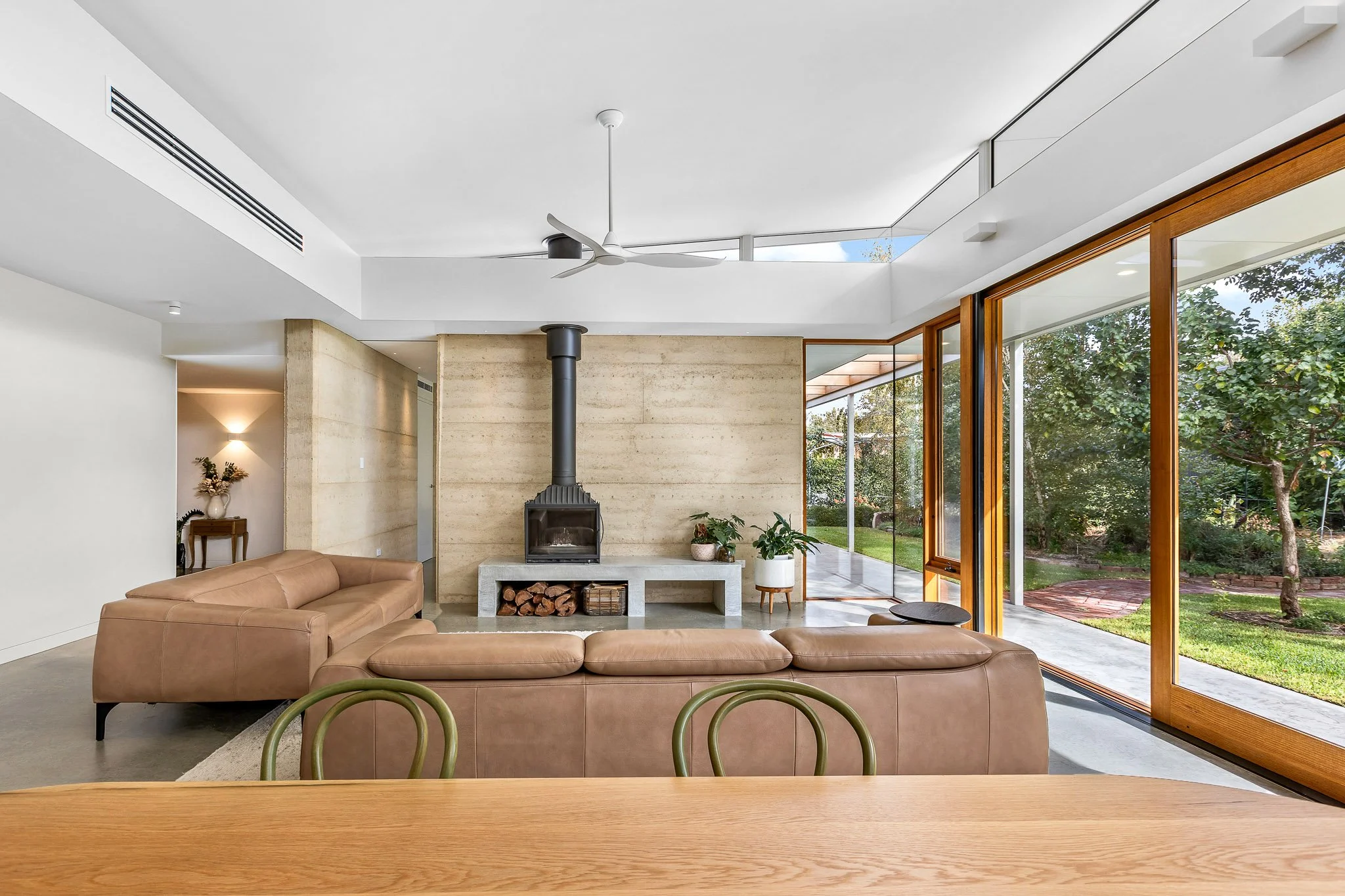 Modern living room with beige sectional sofa, fireplace with stacked wood, large glass windows, indoor plants, and view of a green yard outside.