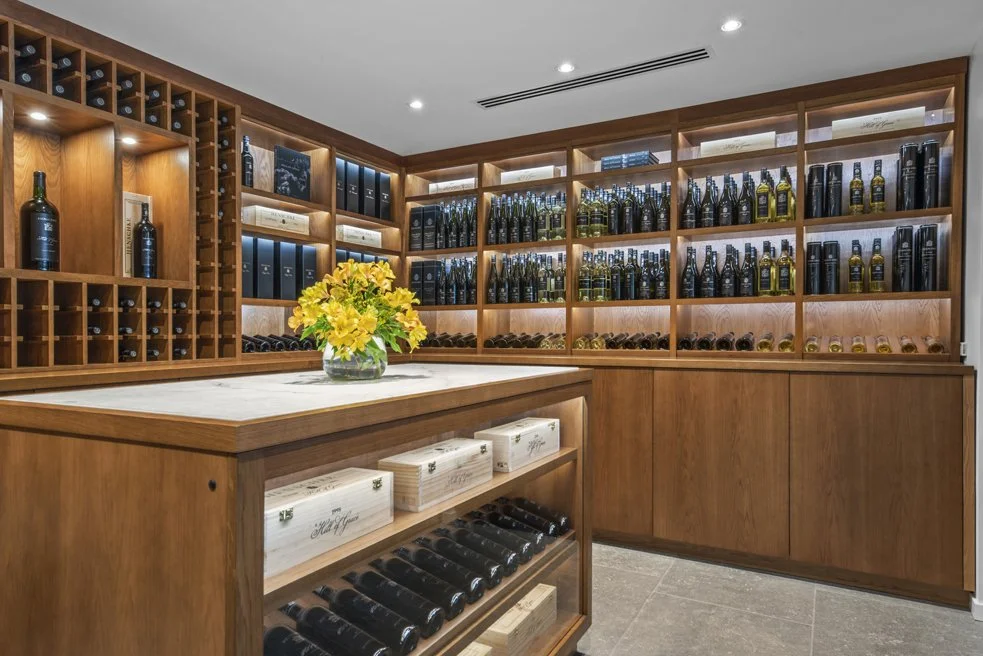 A wine cellar with wooden shelves filled with bottles of wine, wine boxes, and wine accessories. There is a wooden counter with a vase of yellow flowers in the center.