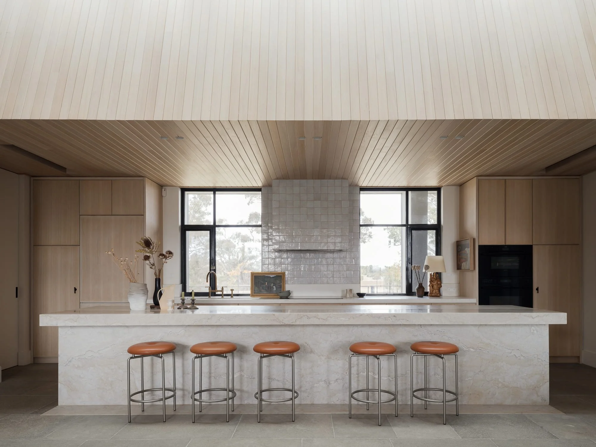 Modern kitchen with marble island, four brown bar stools, light wood cabinetry, large black-framed windows, and a white tiled chimney.