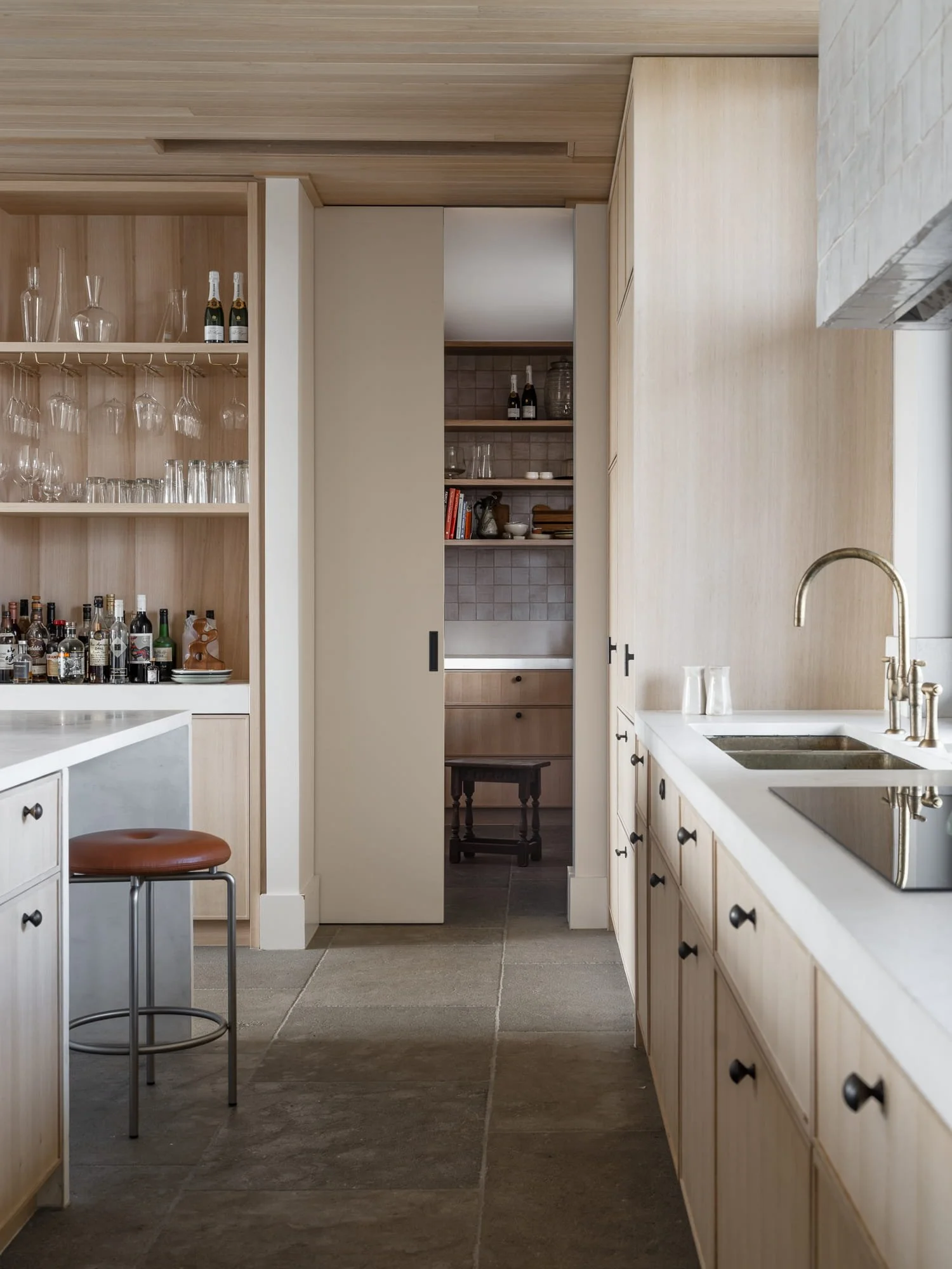 Modern kitchen with light wood cabinetry, a white countertop, double stainless steel sinks, and a brass faucet. Open shelving displays glasses, bottles, books, and decorative items. A small brown stool with a round leather seat is in the foreground, and a sliding door leads to a nook with additional shelves and a small dark wood table.