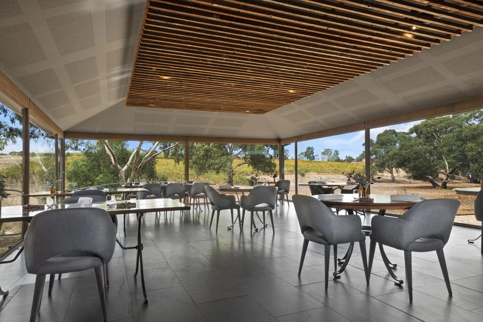 An empty modern restaurant with large glass windows, wooden ceiling panels, gray chairs, and round tables set with glassware, overlooking a natural outdoor landscape with trees and open sky.