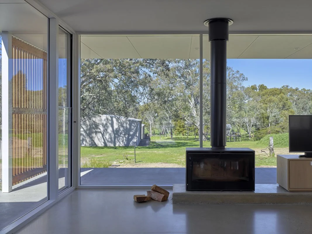 Modern living room with glass sliding doors opening to a green outdoor landscape with trees, a shed, and a clear blue sky, featuring a black fireplace and a small TV.