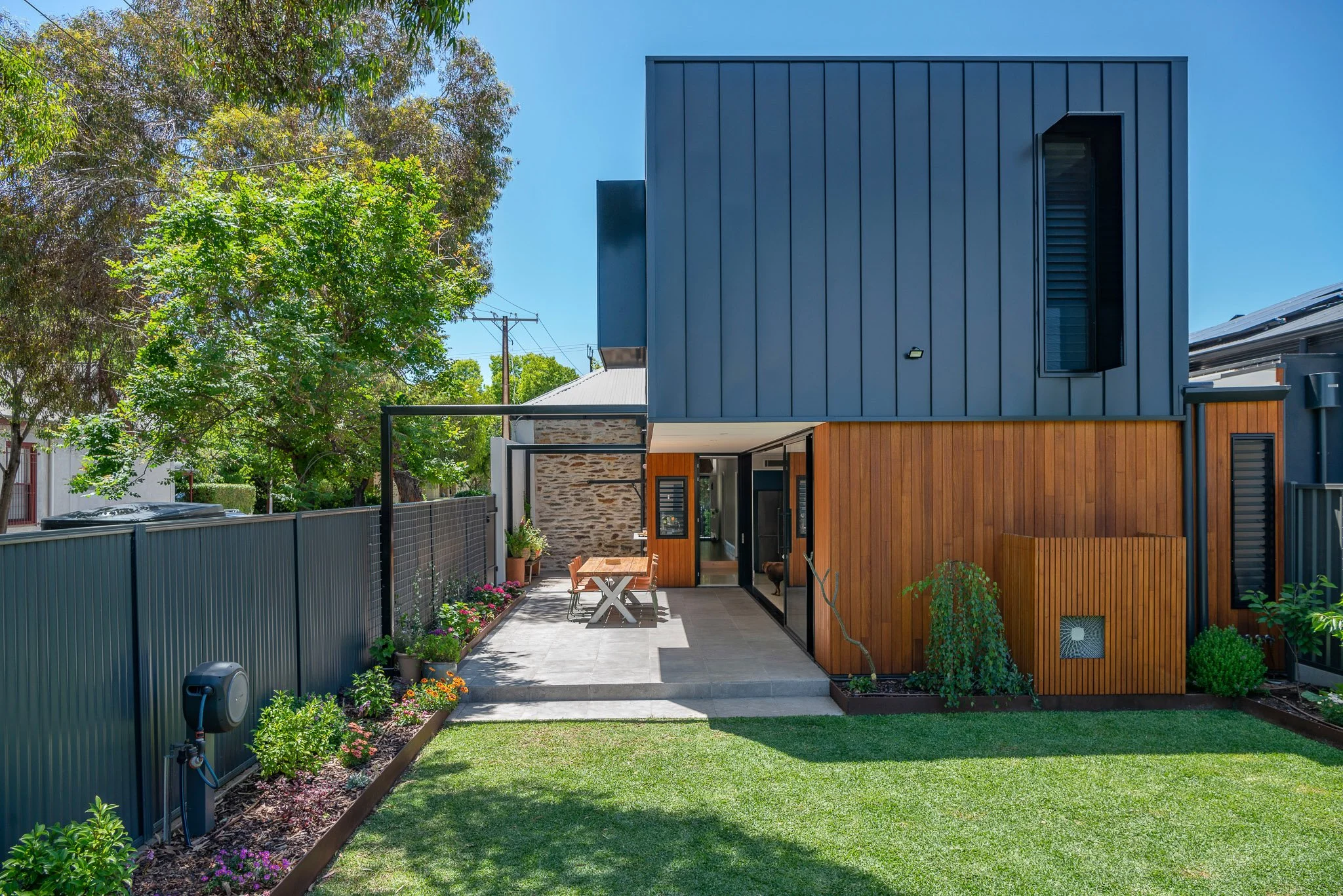 Modern house with a black upper facade and wooden lower exterior, small patio with table and chairs, garden with flowers, green lawn, trees, blue sky, and power lines.