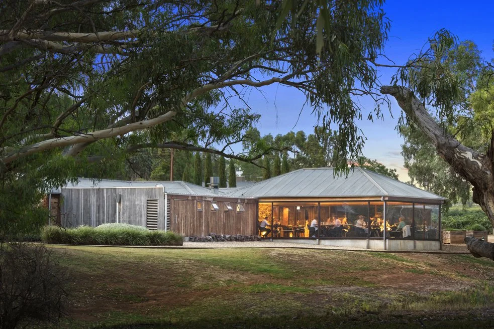 A modern house with a screened-in porch glowing warmly at dusk, surrounded by trees and greenery.