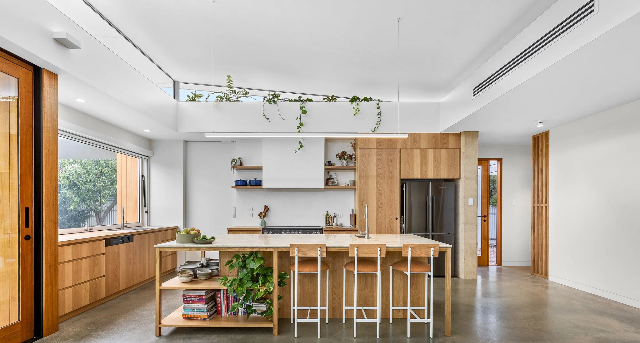 Modern kitchen with wooden cabinetry, a large island with seating, a window showing greenery outside, and minimalistic decor.