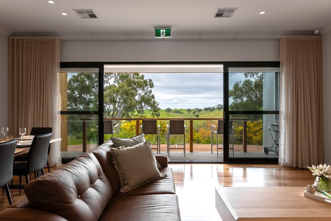 Living room with large glass sliding doors leading to a balcony with chairs, overlooking a green landscape with trees and fields.