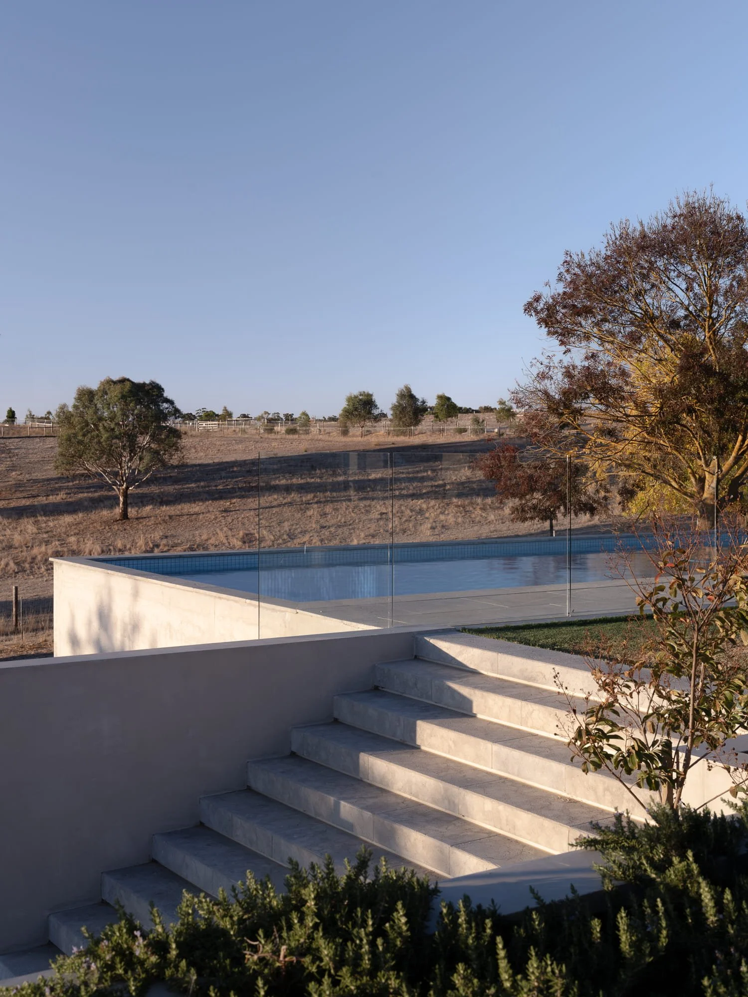 A backyard with a modern concrete pool, beige stairs, and a view of a sloped dry landscape with trees and a clear blue sky.