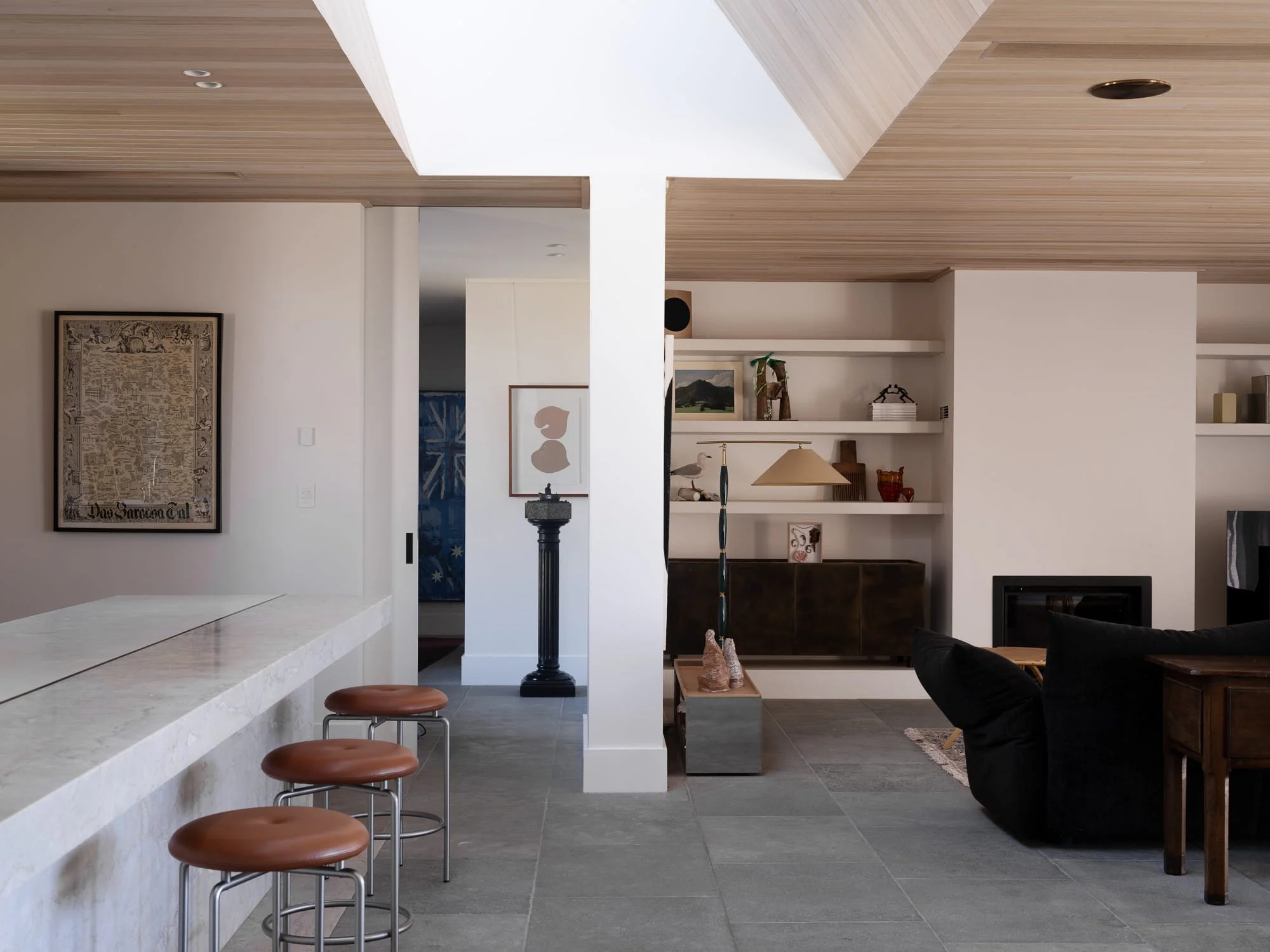 Interior view of a modern living space with a white marble bar counter and four brown stools. The background shows a white wall with artworks and built-in shelves decorated with books and decorative items. The ceiling is made of wood planks with recessed lighting.