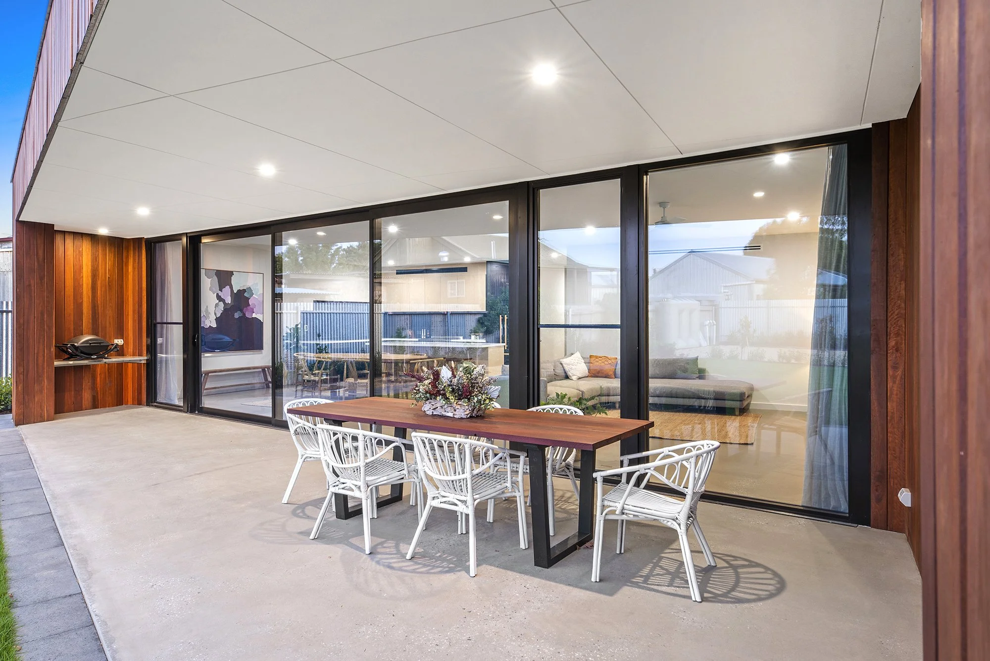 Outdoor patio area with a wooden dining table, white chairs, and a flower arrangement, adjacent to a modern house with large glass sliding doors and an interior living space visible inside.
