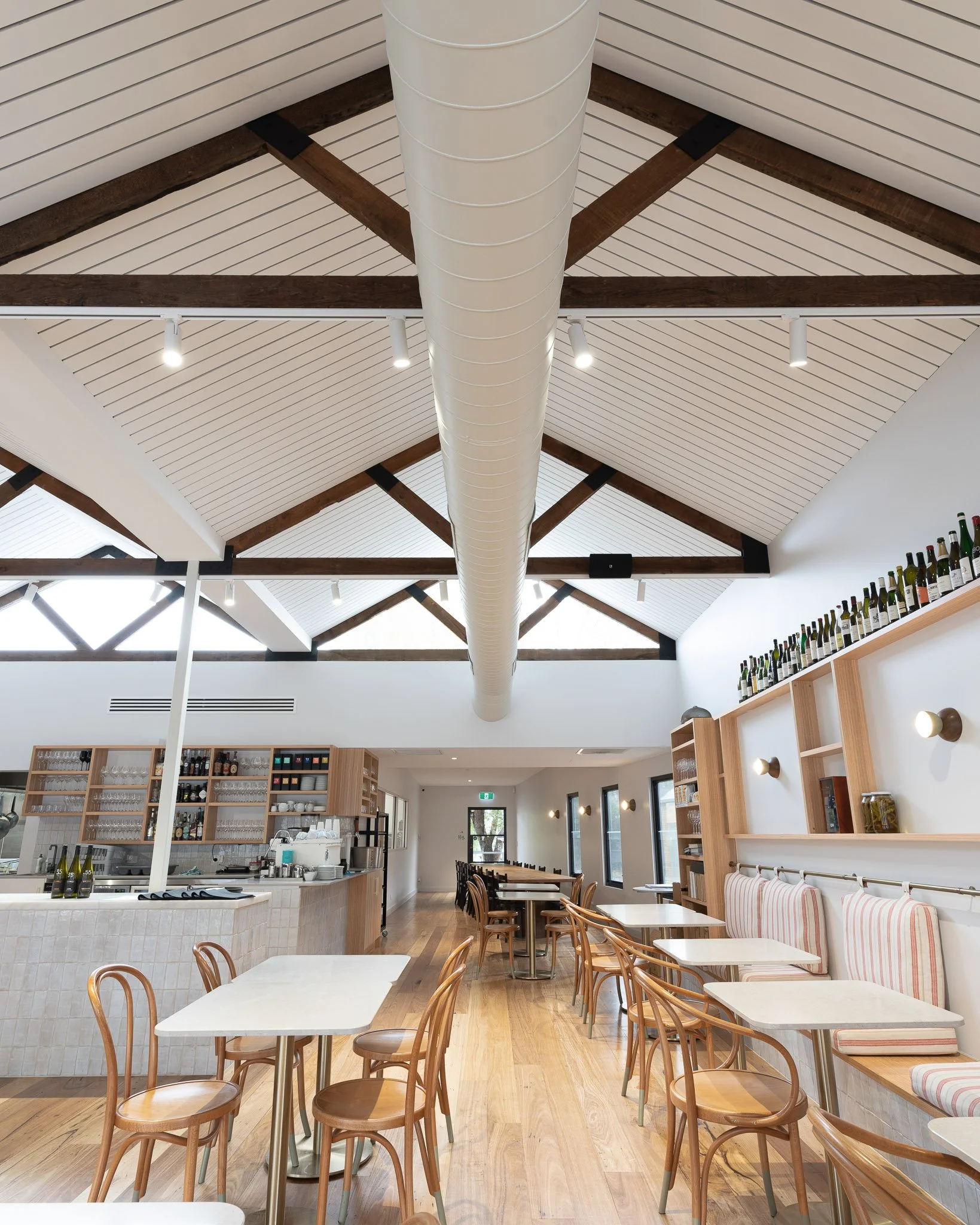 Interior view of a modern cafe or restaurant with wooden chairs and tables, a bar area with glasses and bottles, wood shelving, a white ceiling with wooden beams, and a large white ceiling duct.
