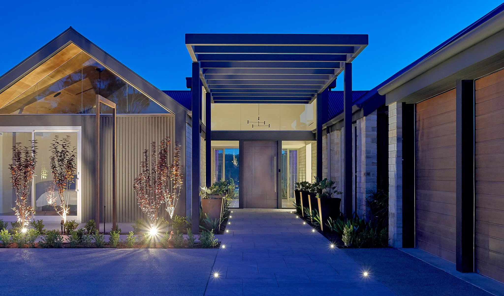 Modern house entrance illuminated with landscape lighting at dusk, featuring a walkway, plants, and architectural design elements.