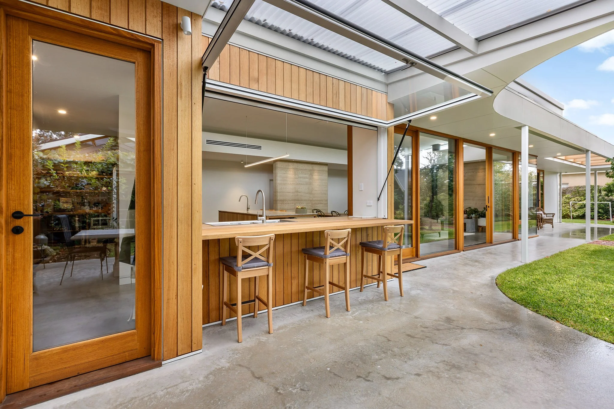 Modern house with wood and glass exterior, open sliding glass door leading to outdoor patio with bar-height wooden seating, view of lush green lawn and trees in the background, under a partially covered patio with a solar skylight roof panel.