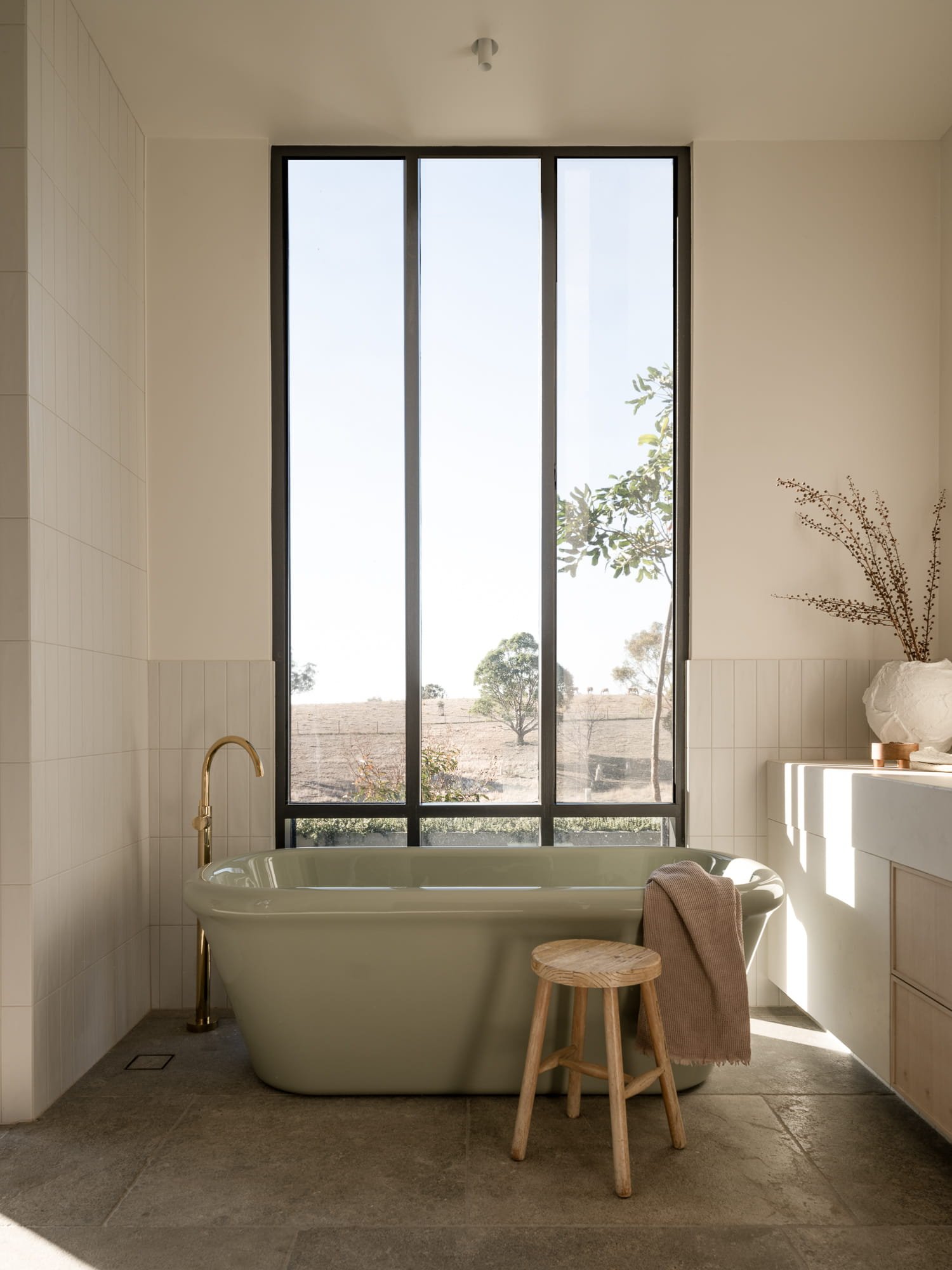 Bathroom with a large window overlooking an outdoor landscape, a clawfoot bathtub, a small wooden stool, and dried decor on a side table.