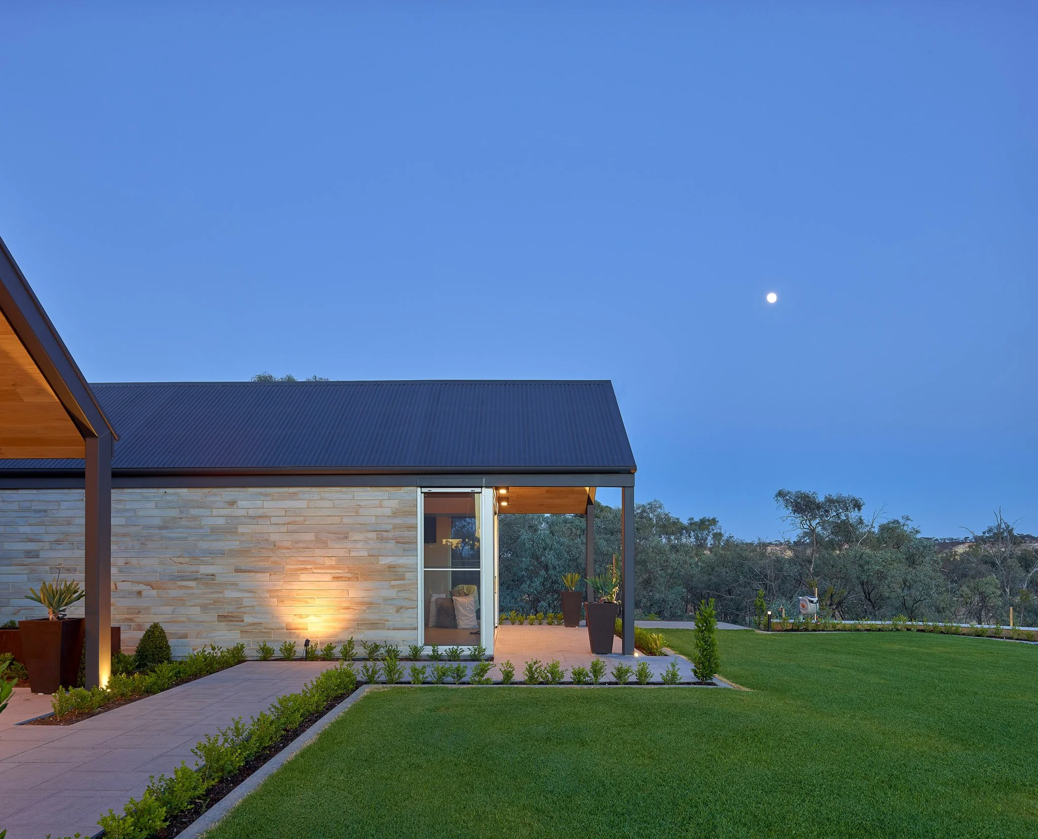 Modern house with stone exterior, large glass window, and patio lights, surrounded by a well-maintained lawn and garden, under a clear evening sky with the moon visible.