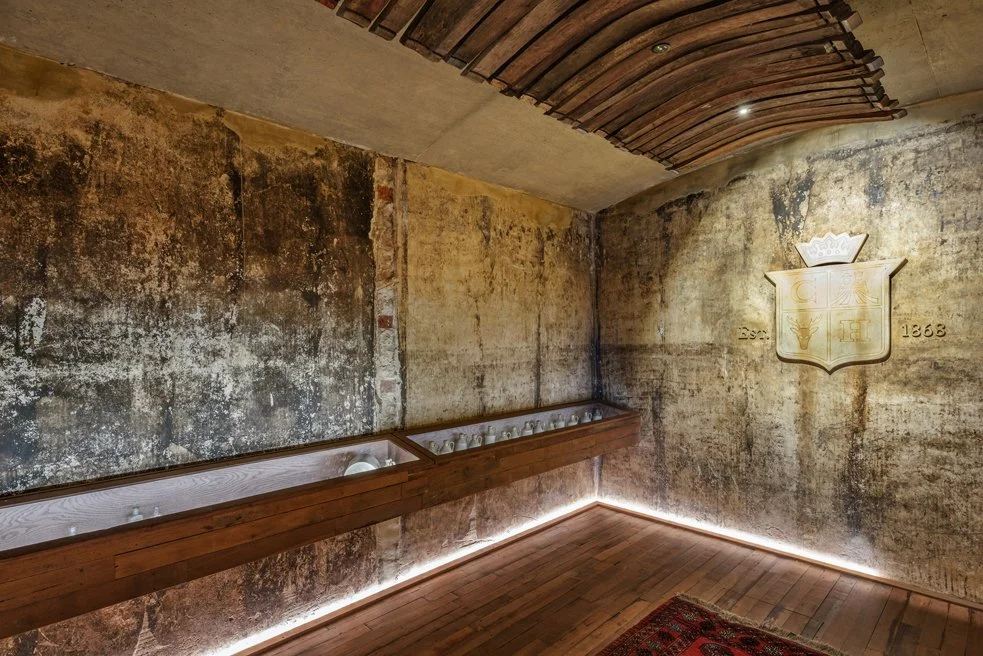 An interior room with distressed, weathered concrete walls and a wooden ceiling. There is a wooden display case along the wall containing small objects. A decorative crest with a crown, shield, and date 1863 is mounted on the wall, with a small rug on the wooden floor and LED lighting along the base of the wall.