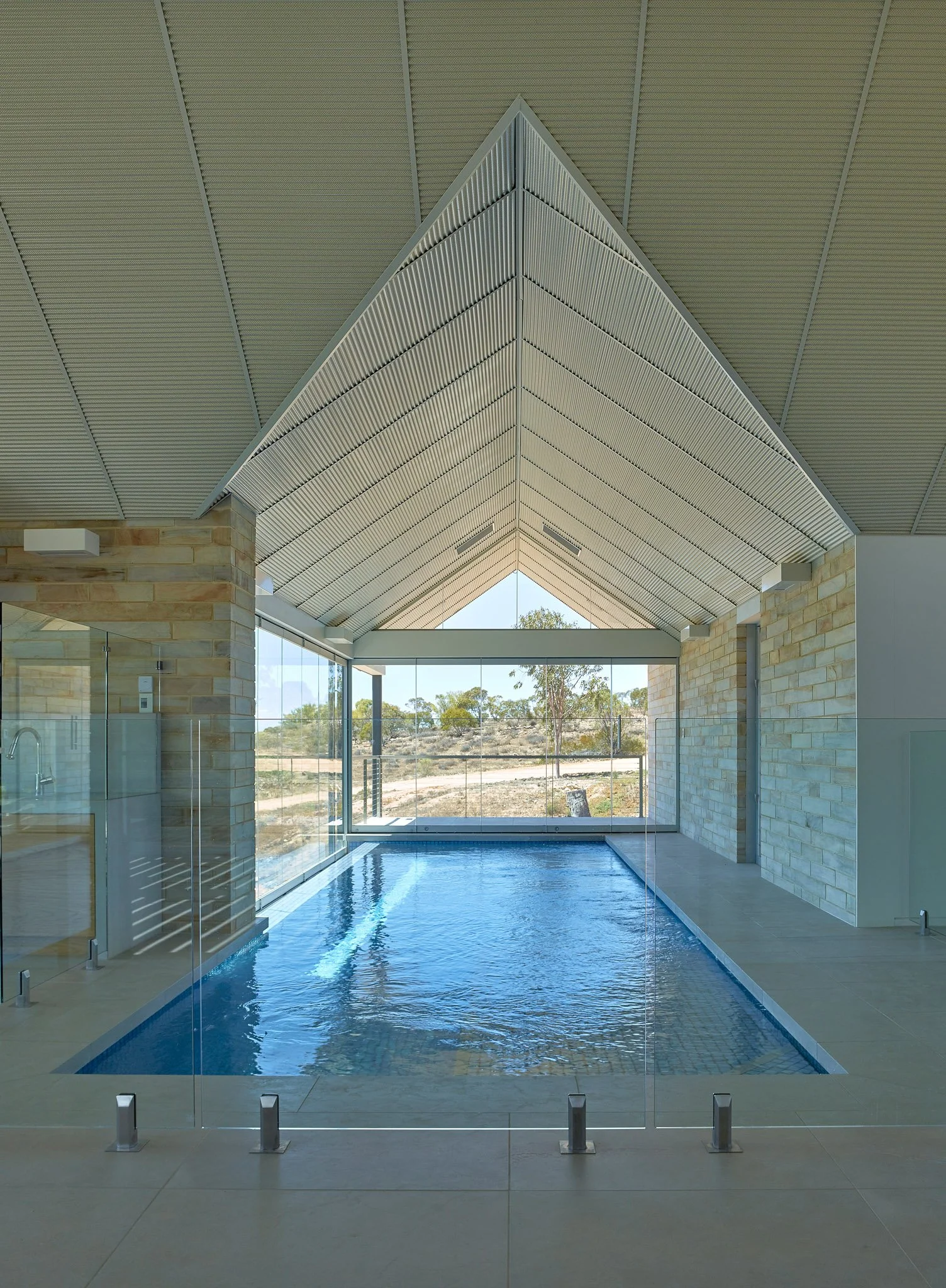 Indoor swimming pool with glass walls, natural light, tiled flooring, and a sloped ceiling with embedded lighting.
