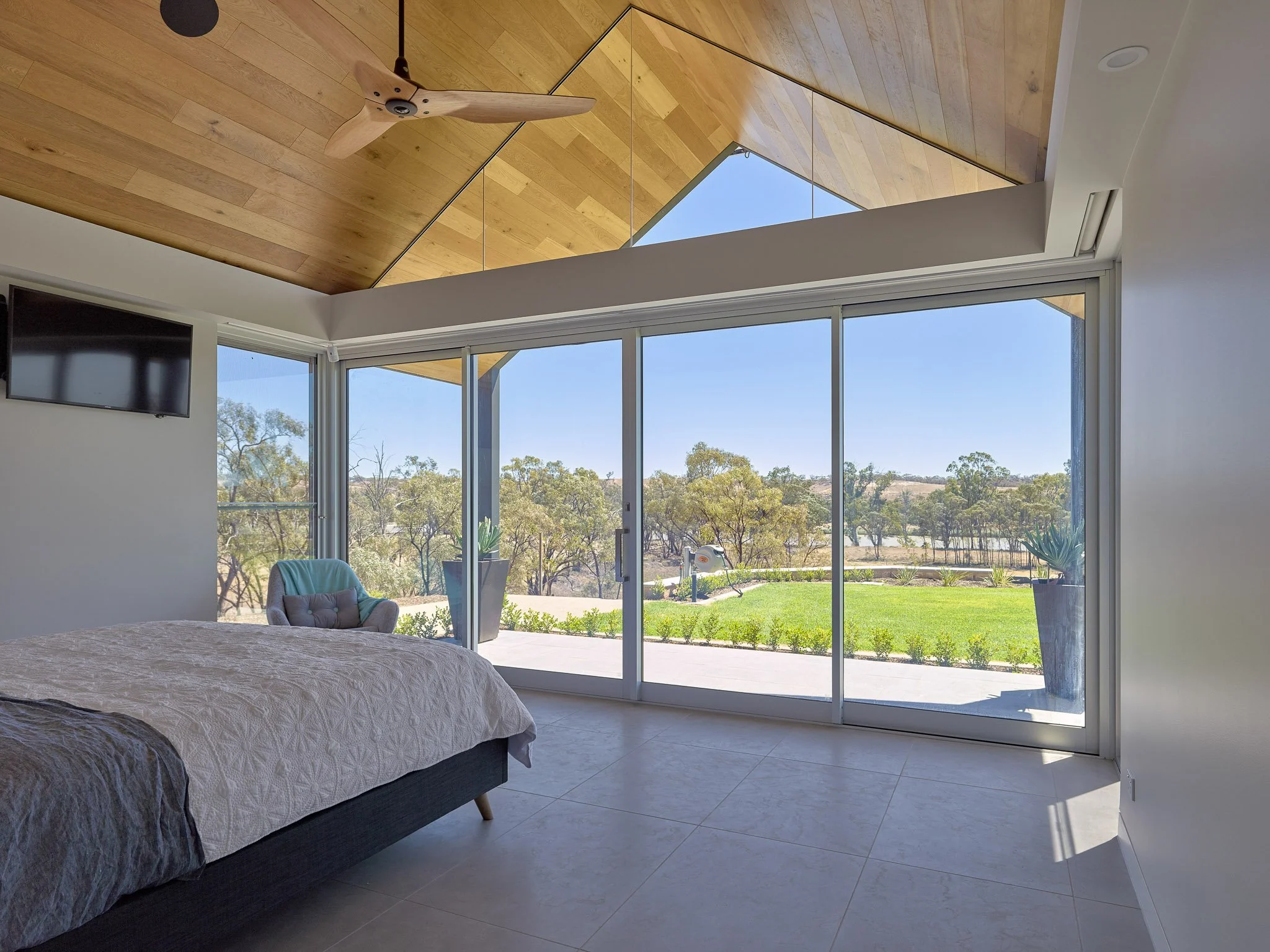 A bedroom with a bed, a TV mounted on the wall, and floor-to-ceiling windows showing an outdoor view of trees and a clear sky.