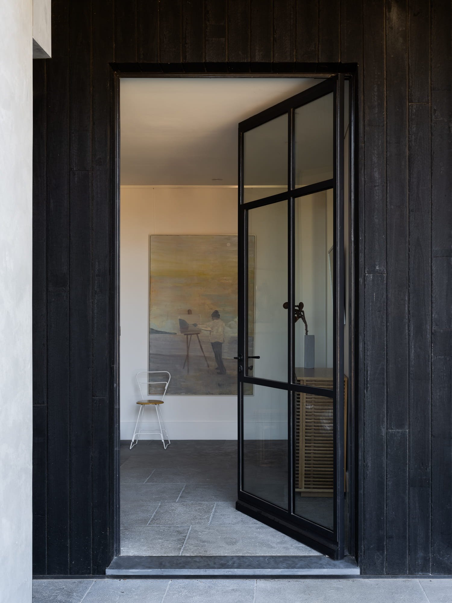 Modern glass door with black frame open to interior with grey tile floor, white walls, a chair, a painting, and a sculpture inside.