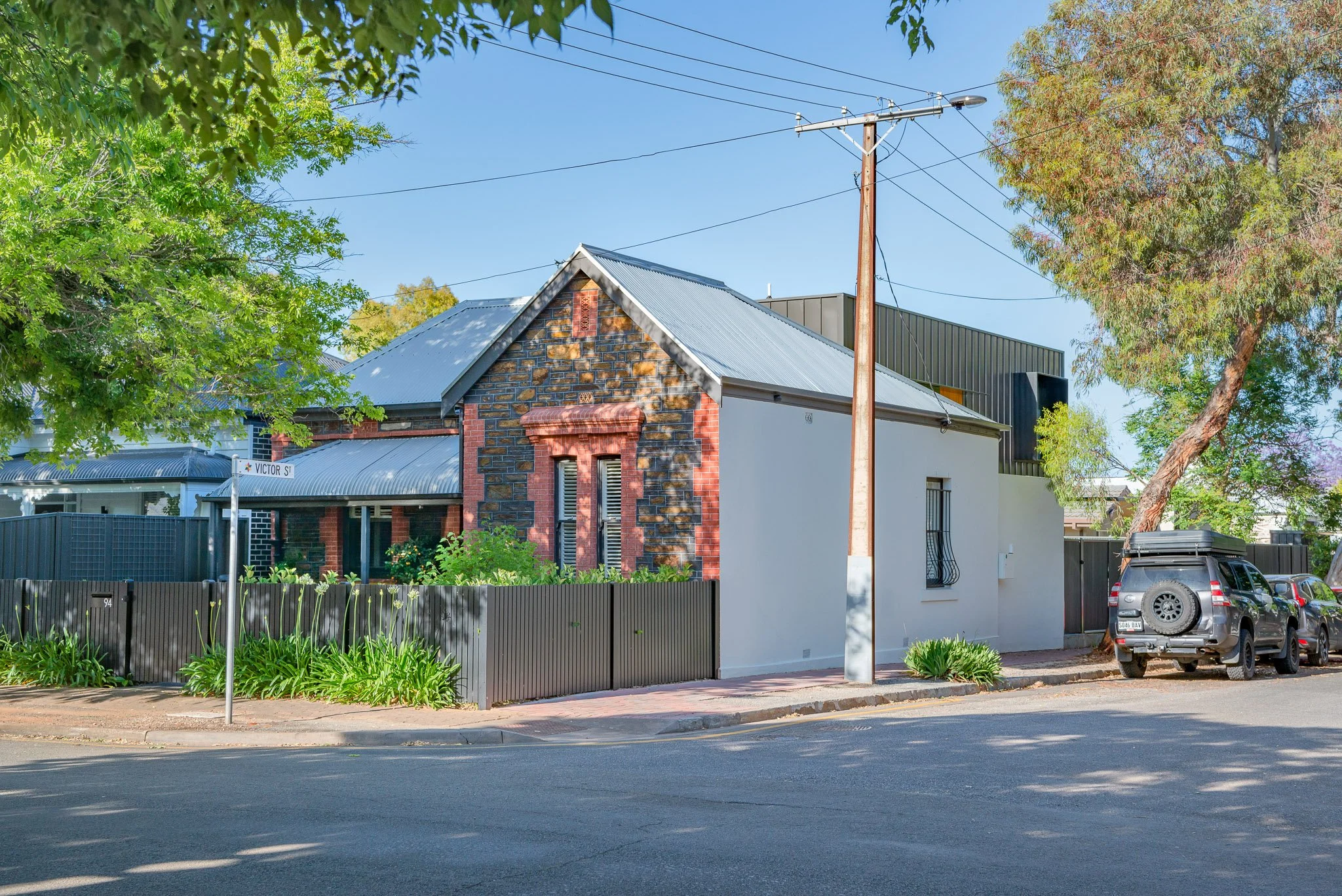 A residential area with a brick house that has a grey metal roof, surrounded by trees and a black fence. There are cars parked next to the house and a street sign that reads 'Victor St'. A utility pole with power lines is in the foreground, with a clear blue sky overhead.