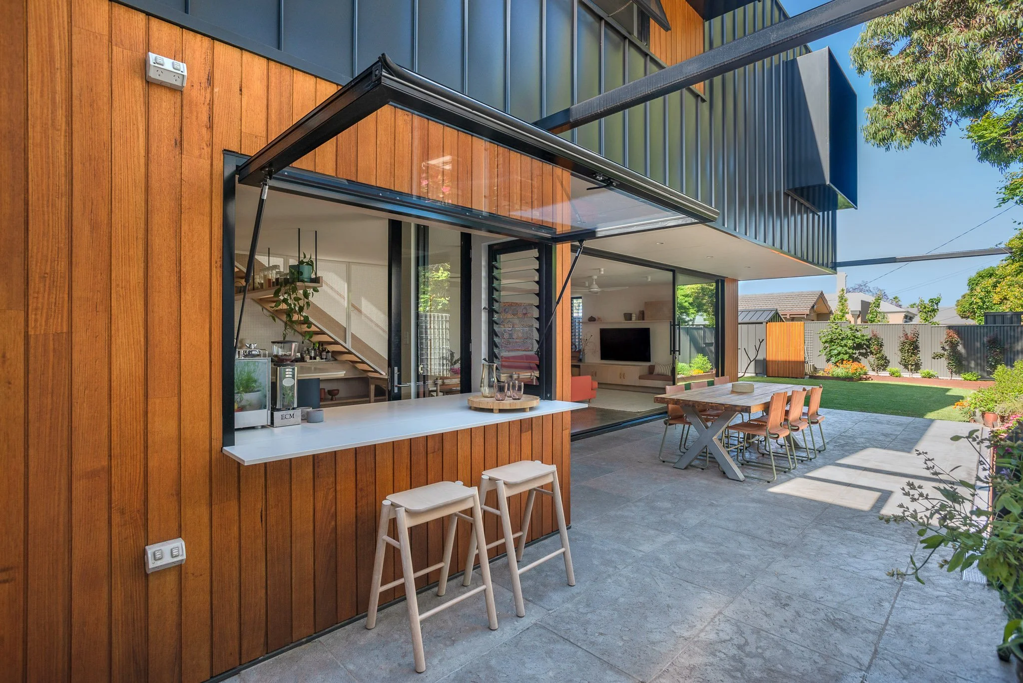 Modern house exterior with large windows, wooden siding, and outdoor seating area including a dining table and chairs, with greenery and a blue sky in the background.