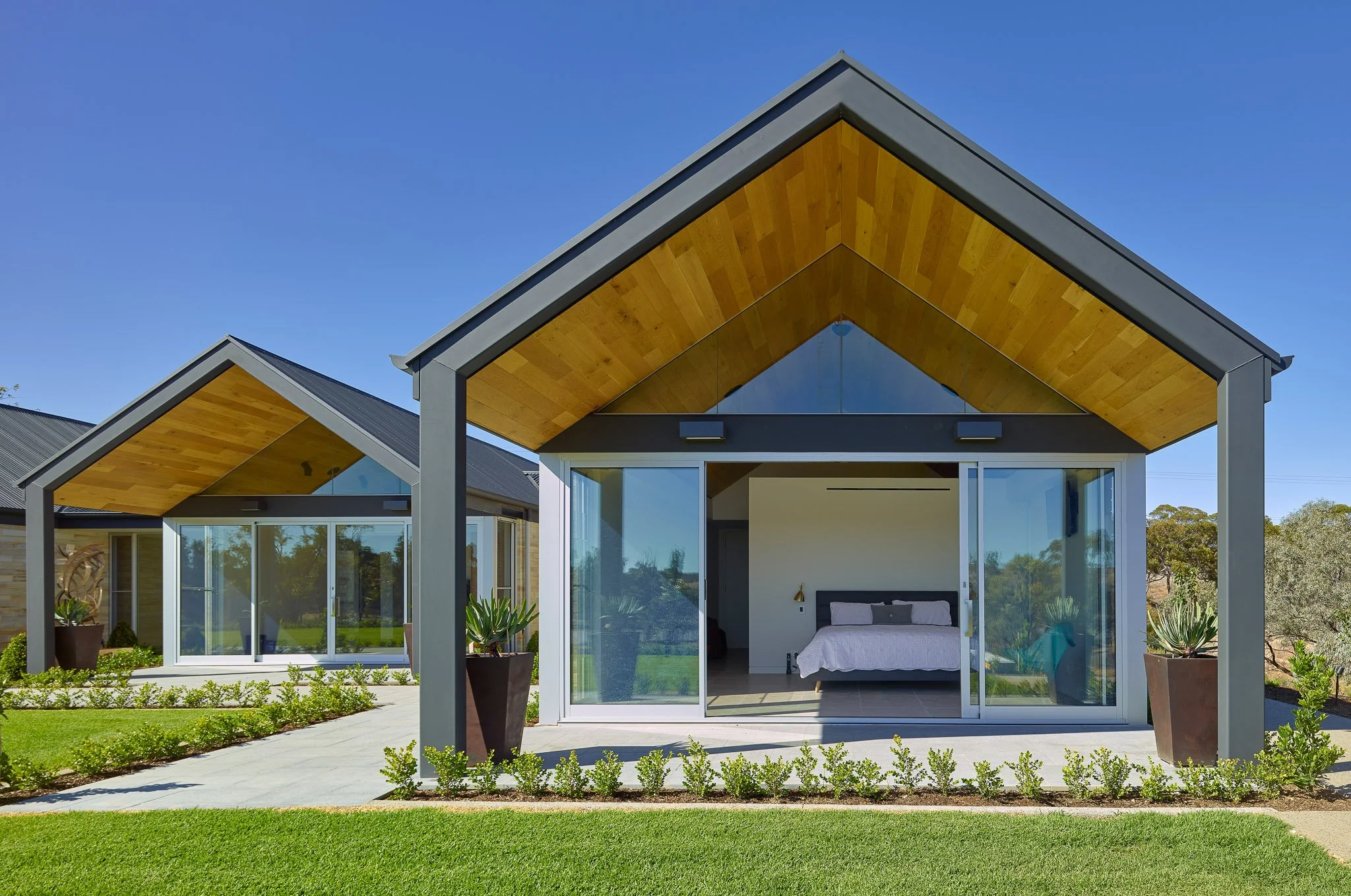 Modern house with large glass windows and sliding doors, featuring a wooden roof extension, surrounded by a well-kept garden with green grass and potted plants, under a bright blue sky.
