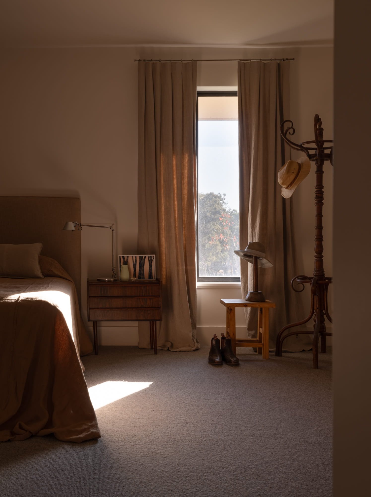 A cozy bedroom with a bed partially visible on the left, a nightstand with table lamp and decorative items, beige curtains on a window letting in natural light, a small wooden stool, a pair of brown boots, a wooden coat rack with a hat, and a pair of eyeglasses, with warm beige carpet flooring.