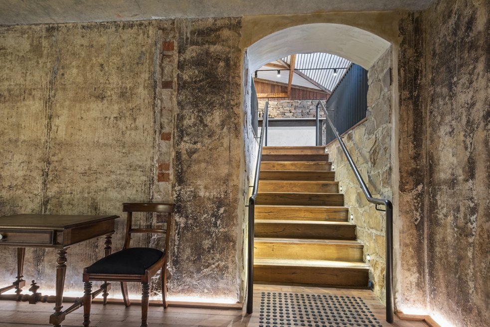 Interior view of a rustic room with weathered walls, an antique wooden table, a matching chair with a black cushion, and a staircase leading to an upper level, visible through an arched doorway.