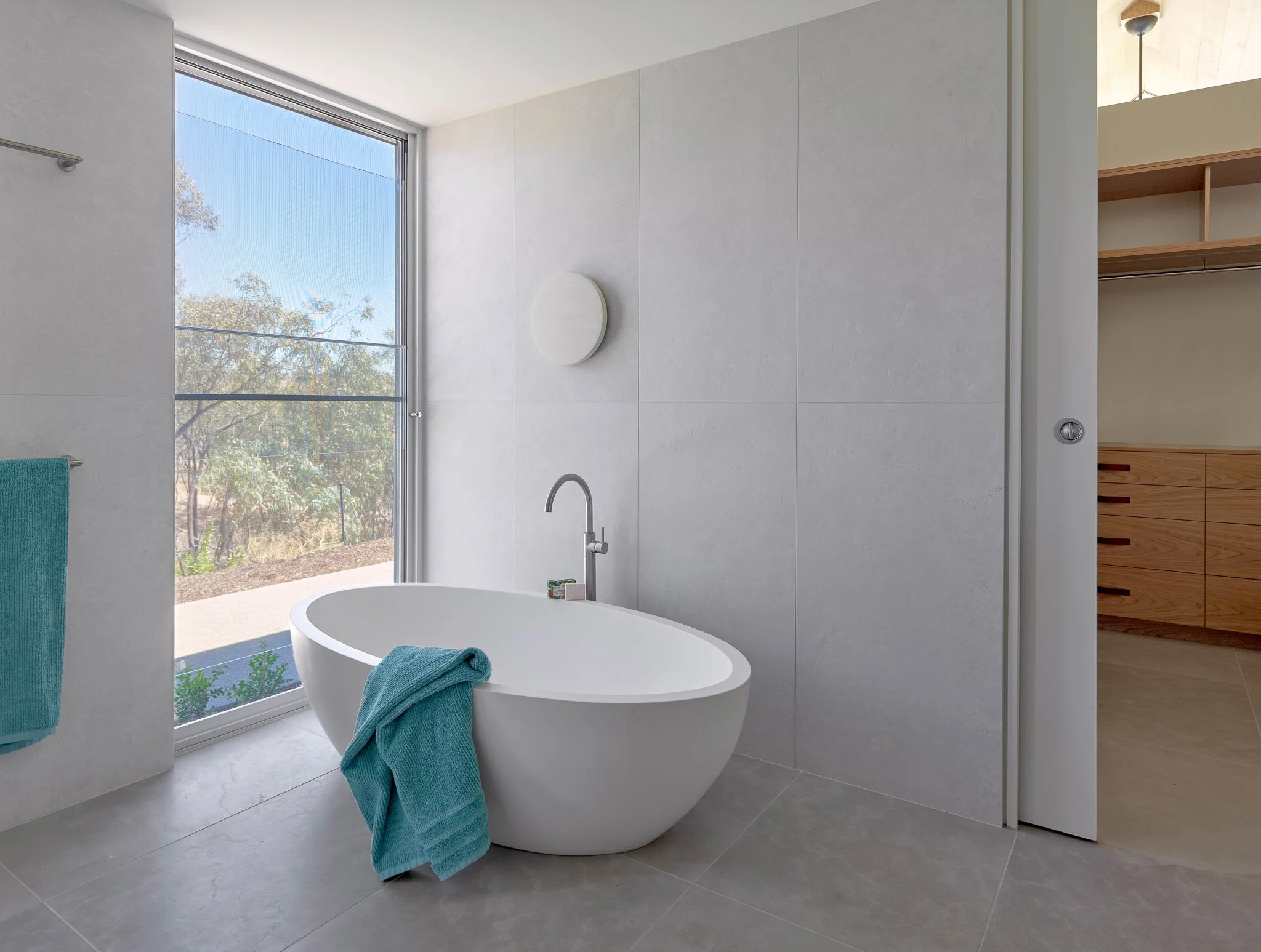 Modern bathroom with a freestanding bathtub near a large window, with teal towels hanging on a railing and a wooden closet.