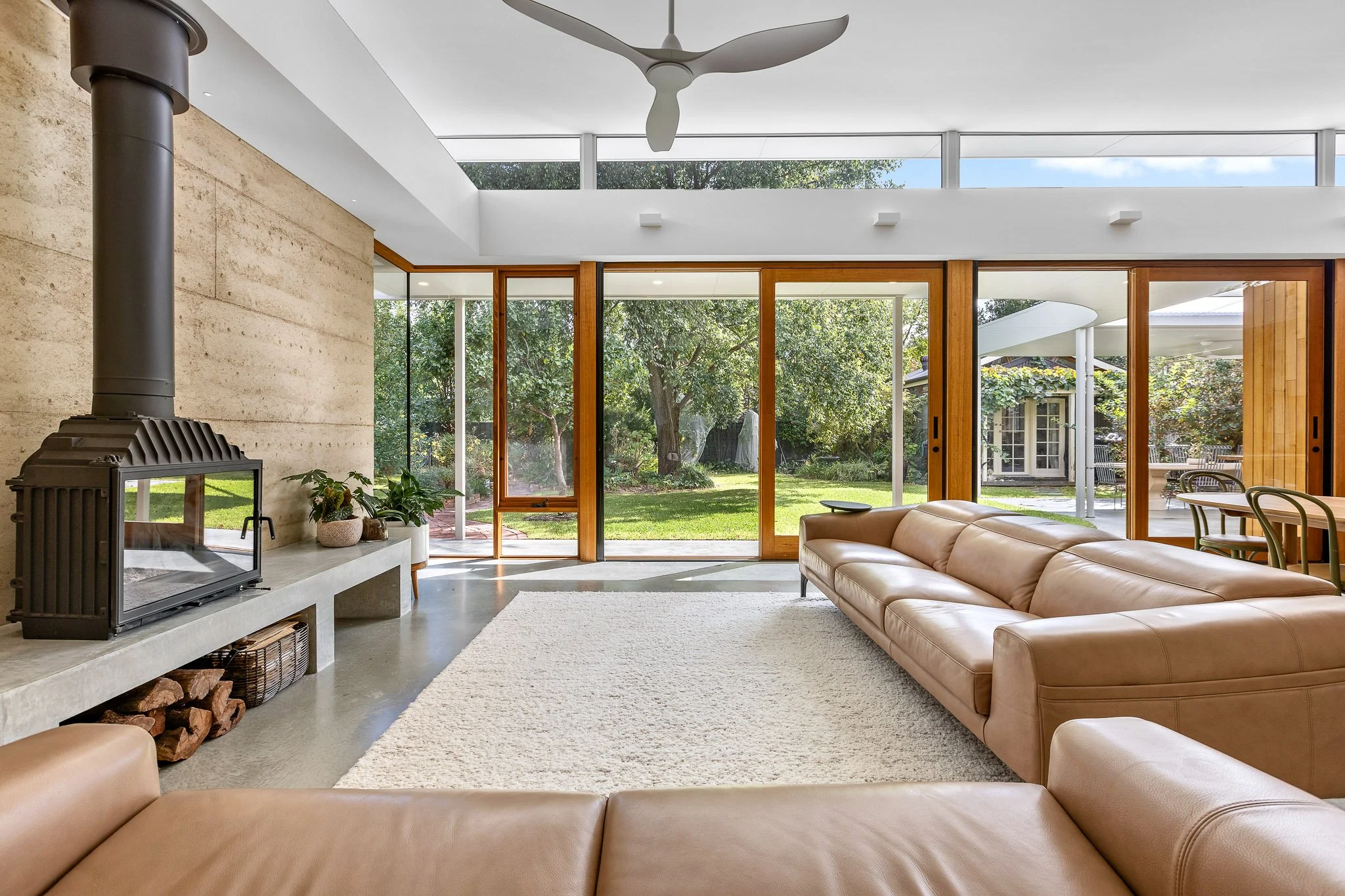 Modern living room with beige leather sectional sofa, wood fireplace, large glass doors opening to a backyard with trees, and a ceiling fan.