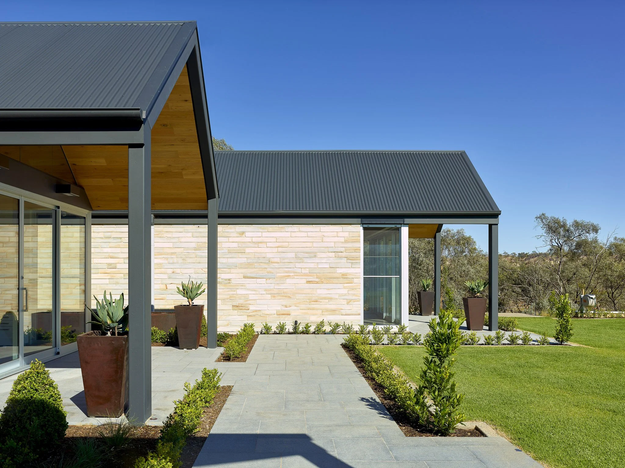 Modern house exterior with metal roof, large windows, landscaped garden, and blue sky.