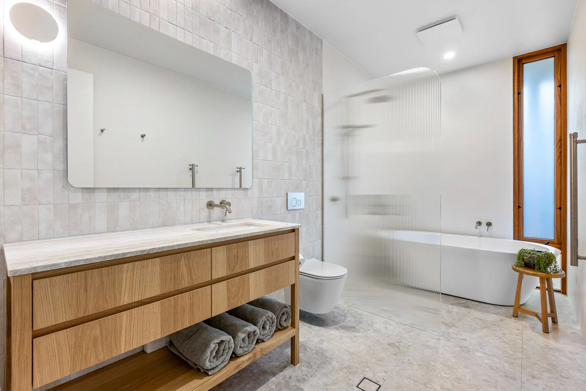 Modern bathroom with white tiled walls, wooden vanity with towel storage, large mirror, toilet, bathtub with frosted glass divider, and a window with wooden frame.