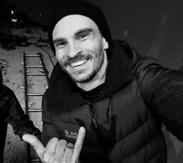 Black and white photo of our DOP smiling man wearing a beanie and jacket, taking a selfie outdoors at night with a long track behind him that was built outdoors.