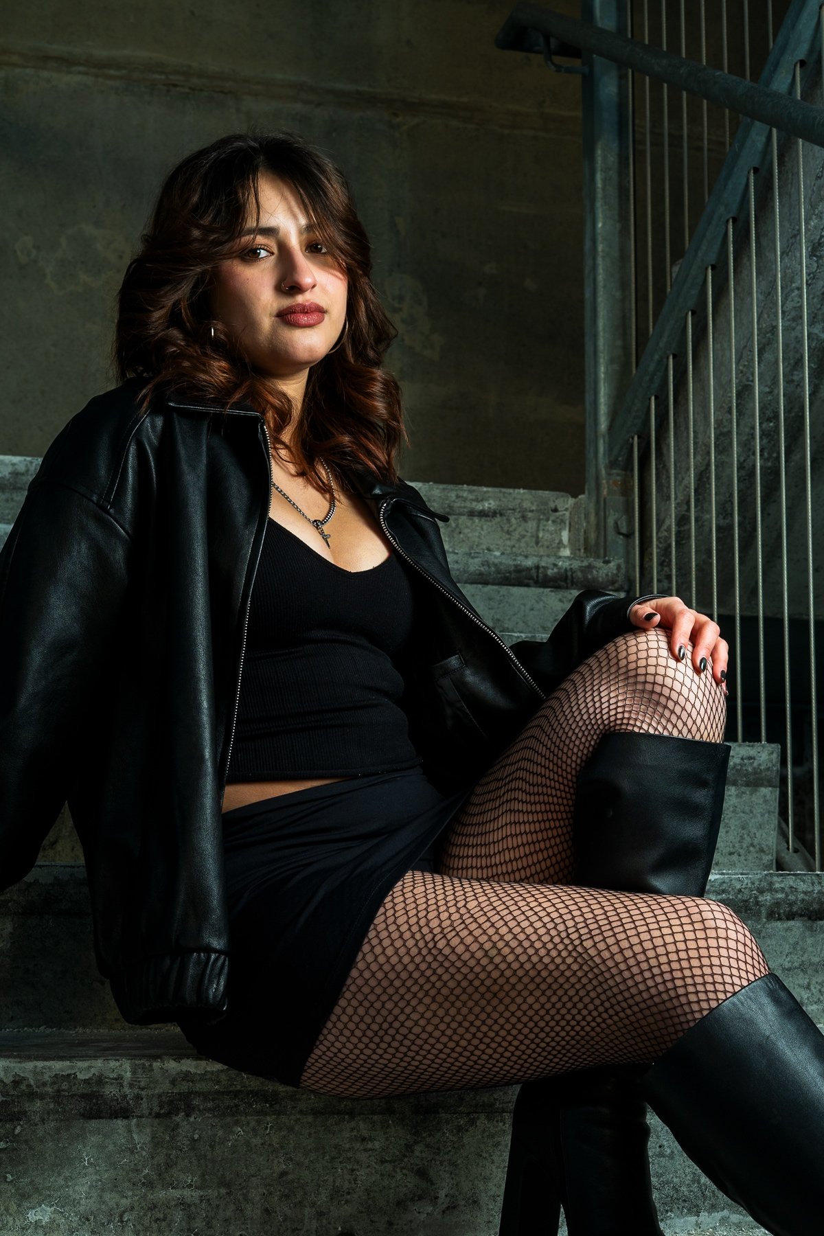 Ariz wearing a leather jacket and fishnets sitting on concrete stairs at Humber College North Campus.