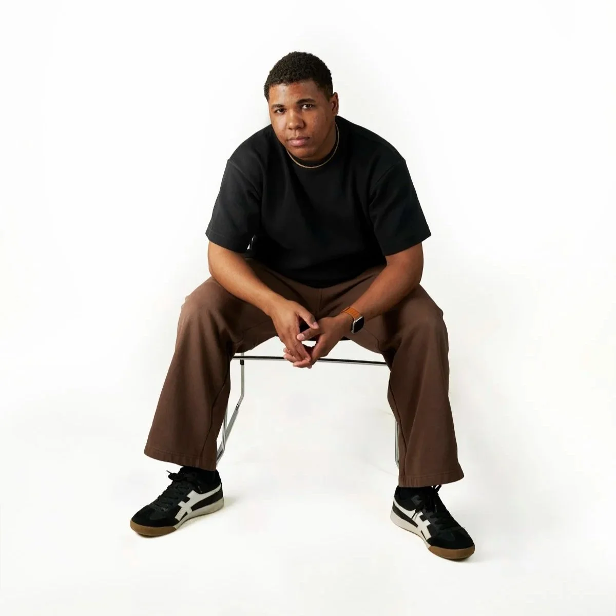 Young man sitting on a metal chair in a studio with a white background, wearing a black t-shirt, brown pants, and black sneakers.