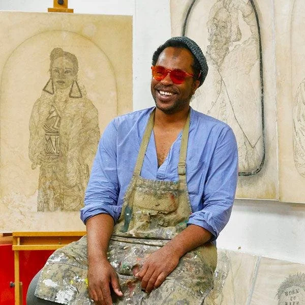 An artist wearing a blue shirt, red sunglasses, and an apron smiling in a studio with drawings of a woman and a map on the wall behind him.