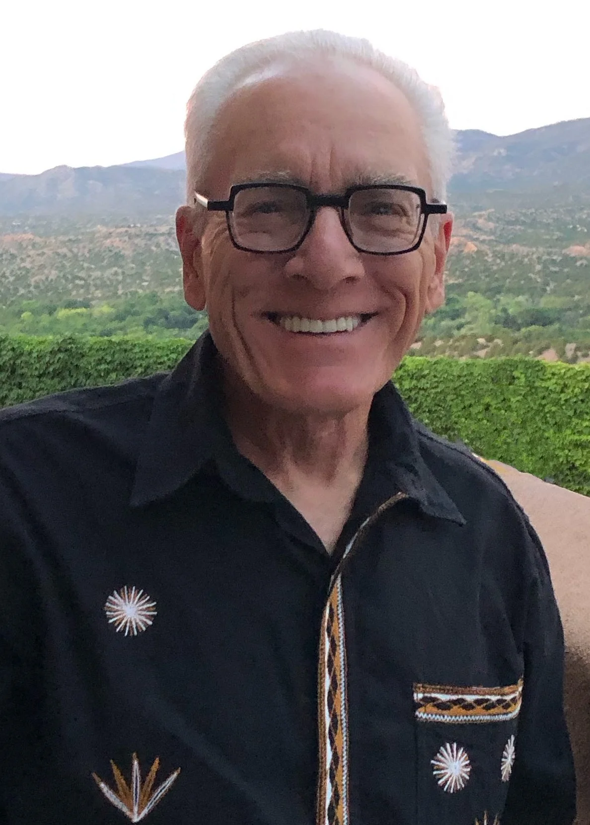 A smiling elderly man with glasses and white hair standing outdoors with mountains and greenery in the background.