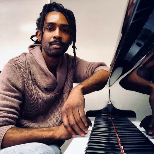 Man with dreadlocks sitting at a grand piano in a room with a plain wall.
