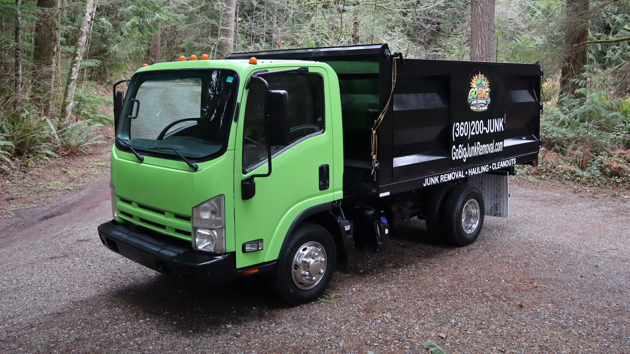 A green and black junk removal truck parked on a forested dirt trail.