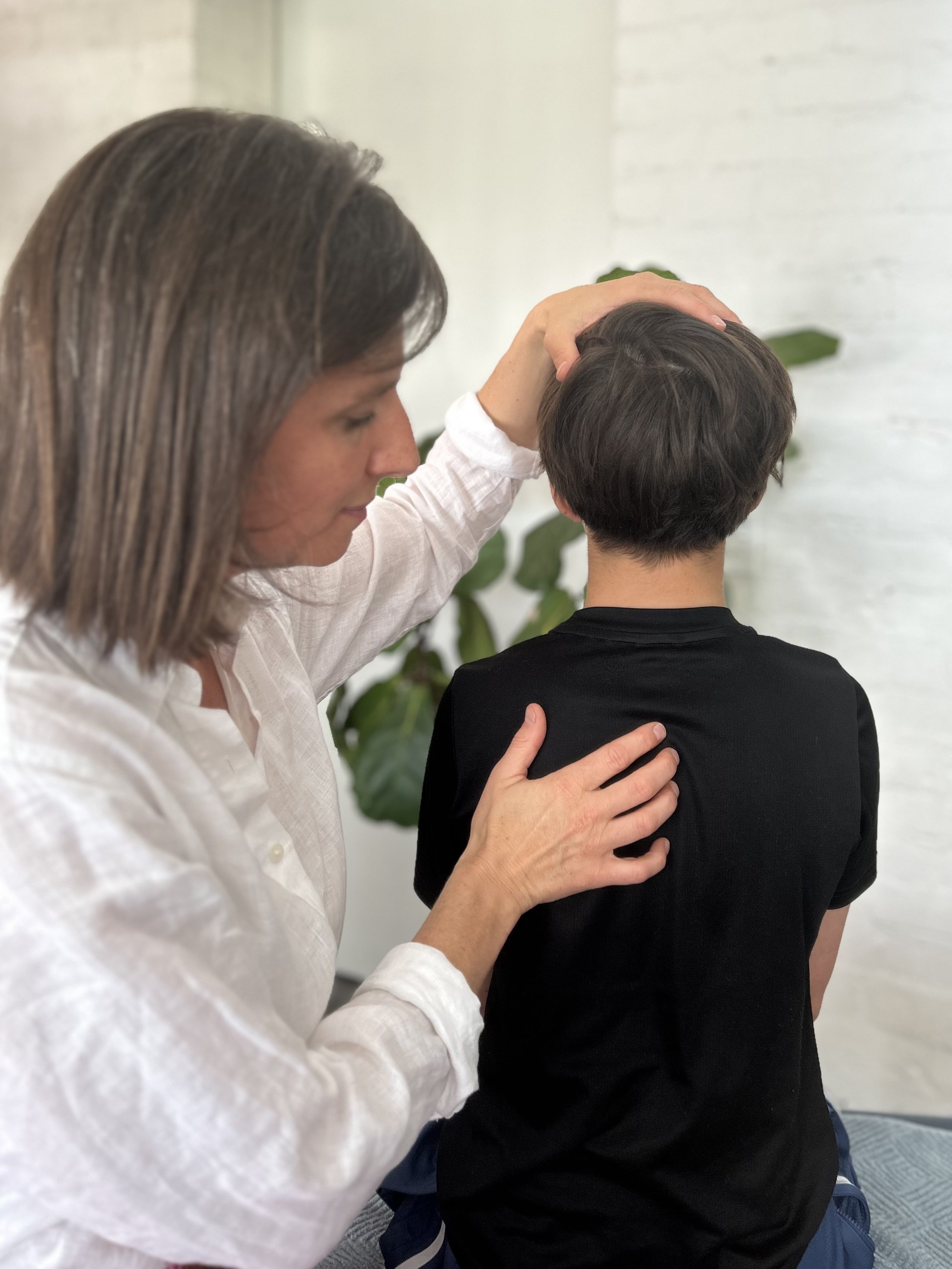 A woman with brown hair in a white shirt gently places her hand on the head of a young boy with dark hair, who is sitting with his back to the camera.