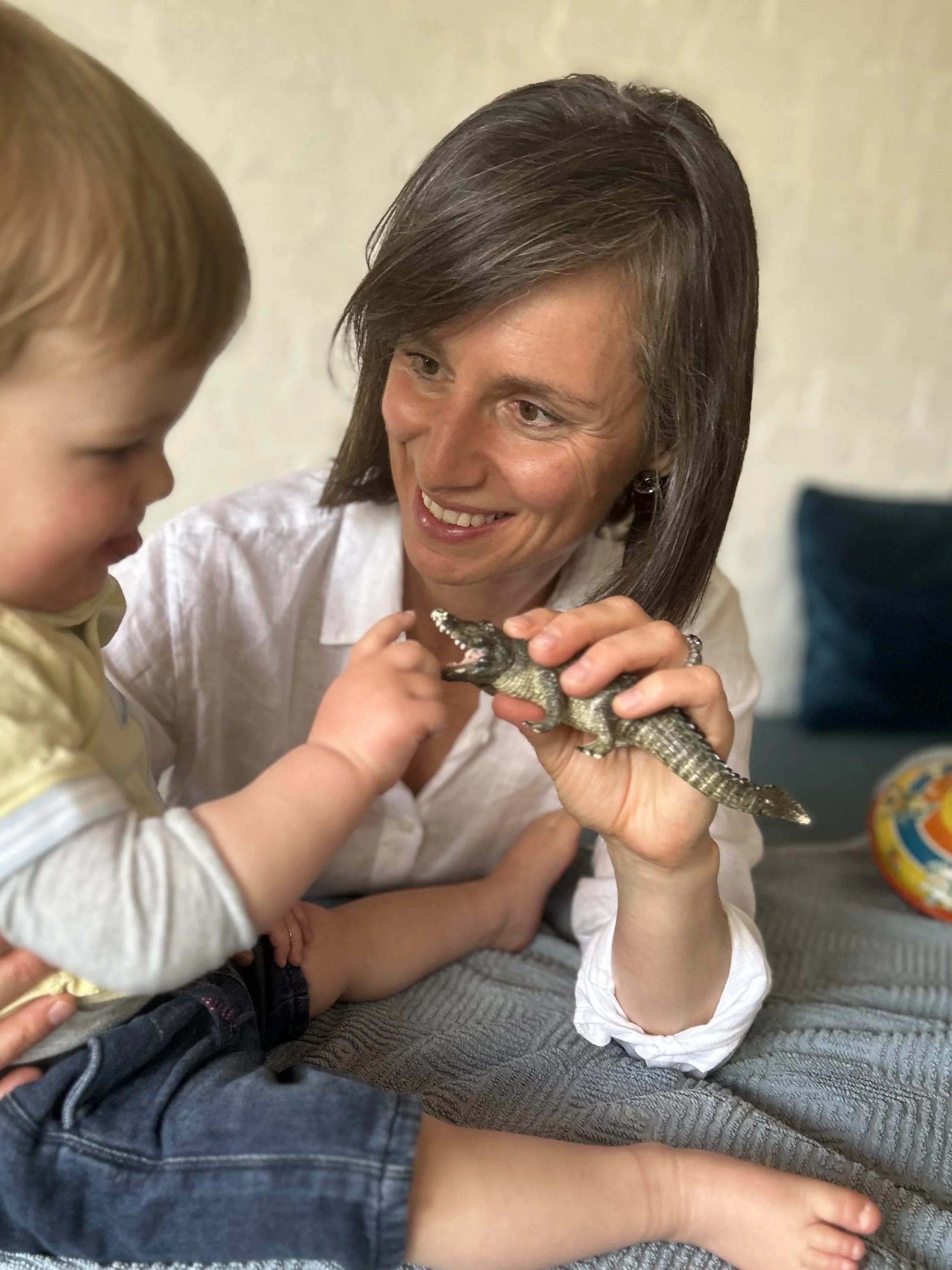 A woman holding a small toy alligator while a young boy touches it, sitting on a gray cushioned surface, with a colorful ball in the background.