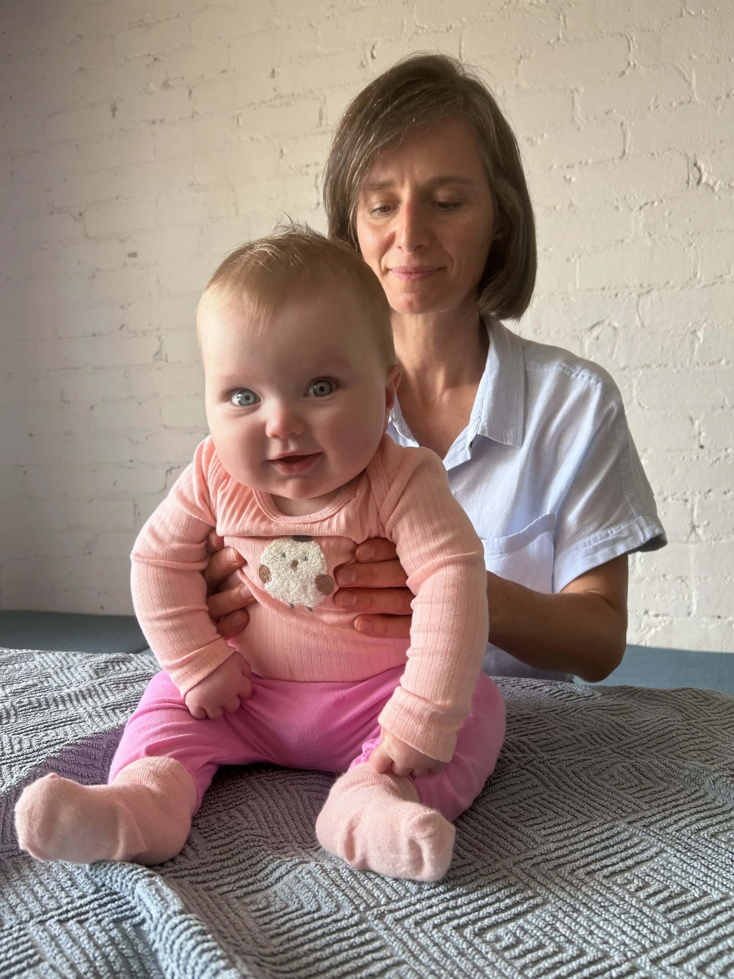 A woman holding a smiling baby girl with big blue eyes, sitting on a textured gray blanket against a white brick wall.