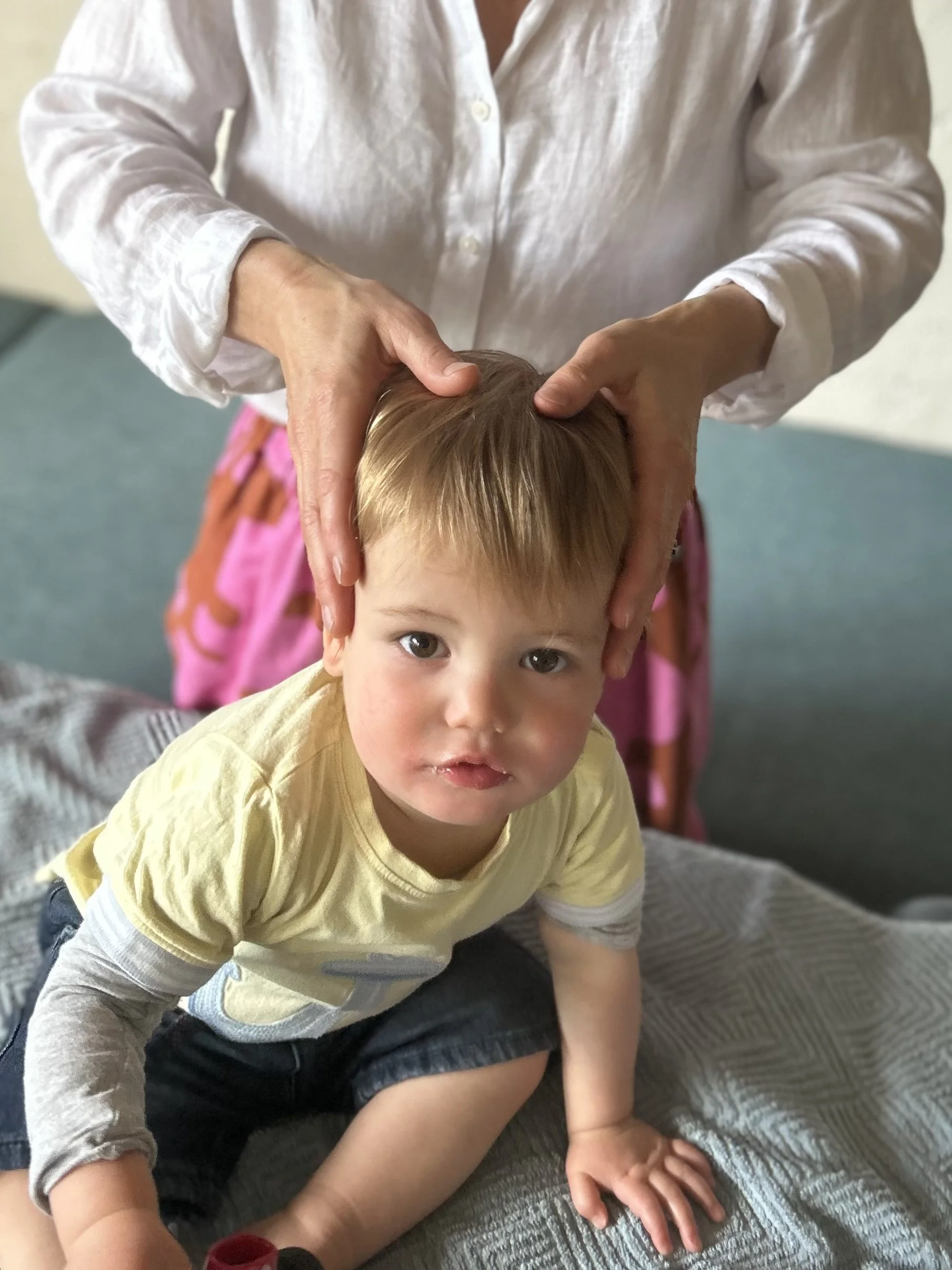 A young boy with light brown hair and blue eyes lying on a bed, looking directly at the camera. An adult, wearing a white button-up shirt, is holding the boy's head with both hands. The bed has a gray textured blanket. In the background, there is a pink and orange backpack.