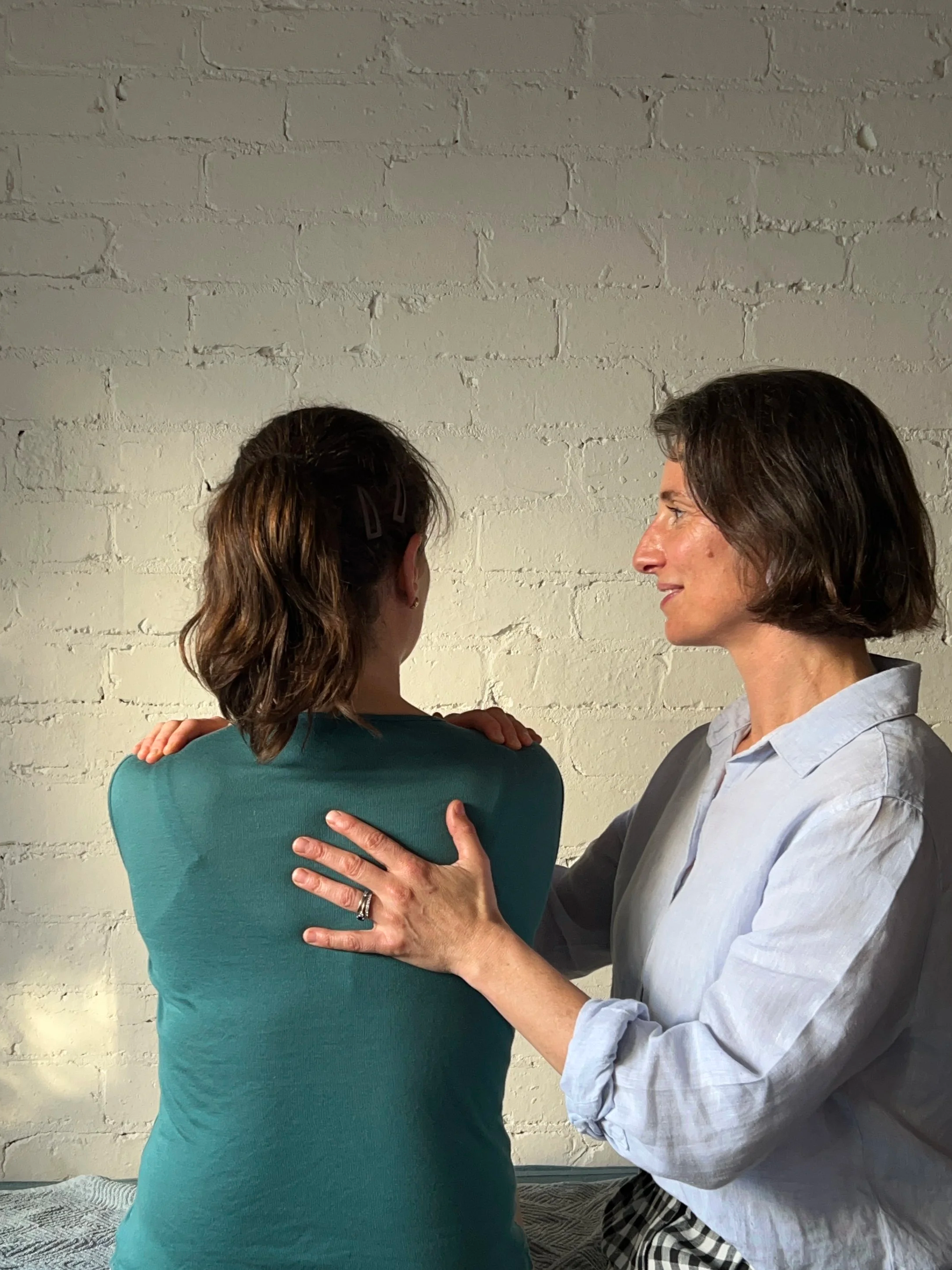 Two women are standing facing each other against a white brick wall. One woman, with shoulder-length brown hair, is wearing a teal shirt, and the other woman, with short dark hair, is wearing a light gray shirt. The woman in gray is touching the shoulder of the woman in teal.
