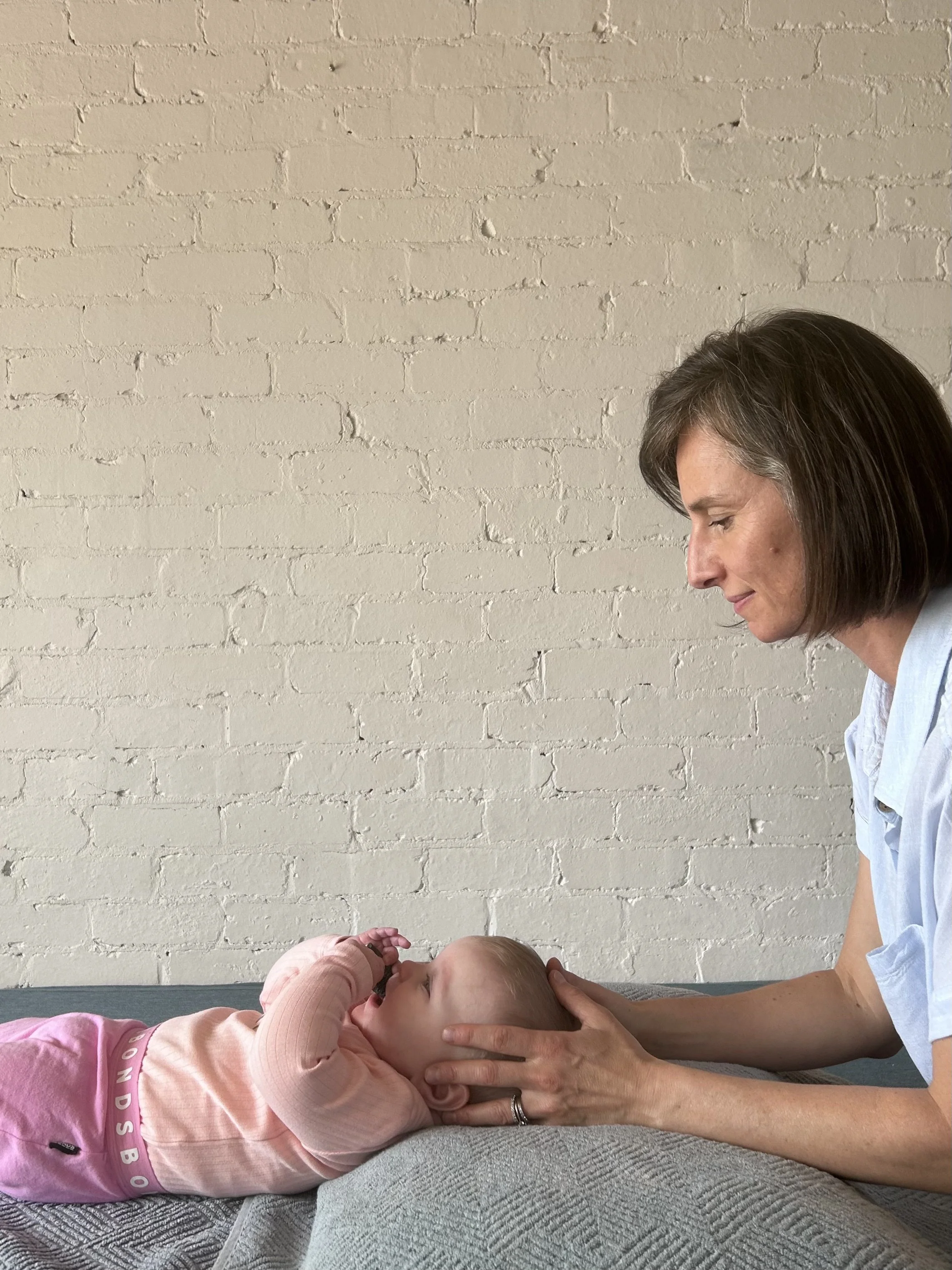 A woman and a young girl lying on their sides facing each other; the woman is gently holding the girl's head and the girl has her hands near her face, with a white brick wall in the background.