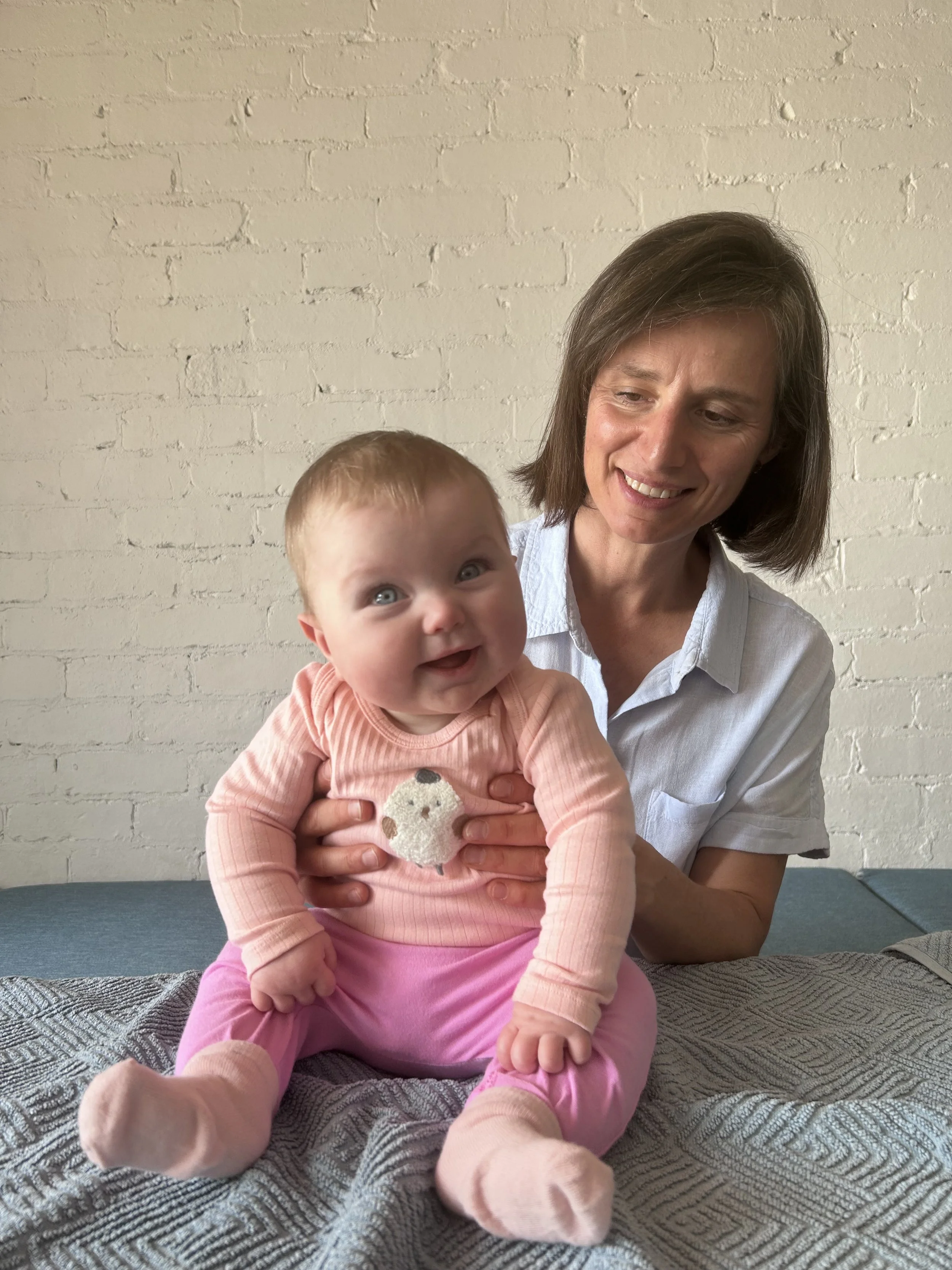 A woman with shoulder-length brown hair smiling at a baby girl with red hair and blue eyes, sitting on a textured gray blanket in front of a white brick wall.